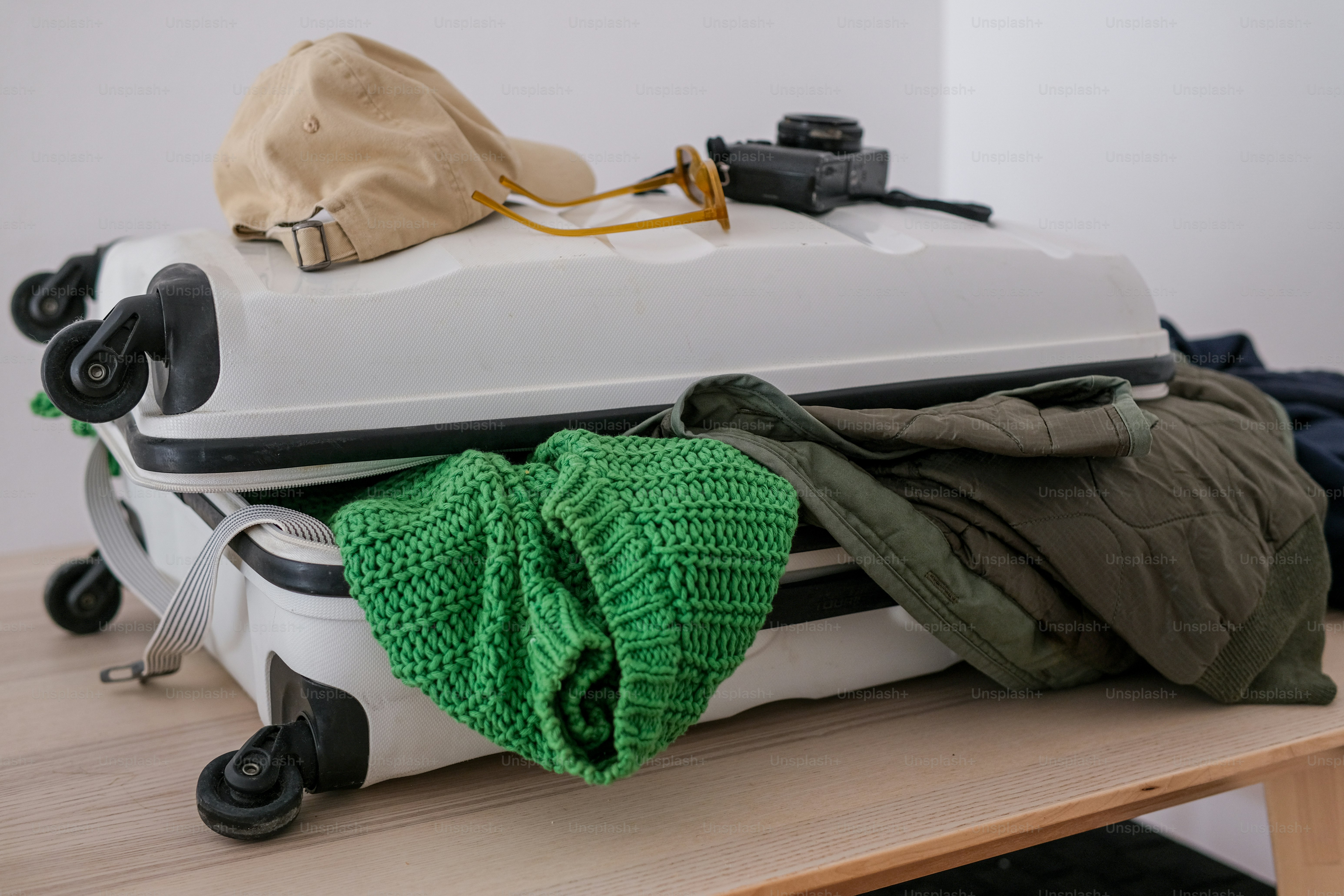 An open white suitcase overflowing with clothes and a cap.