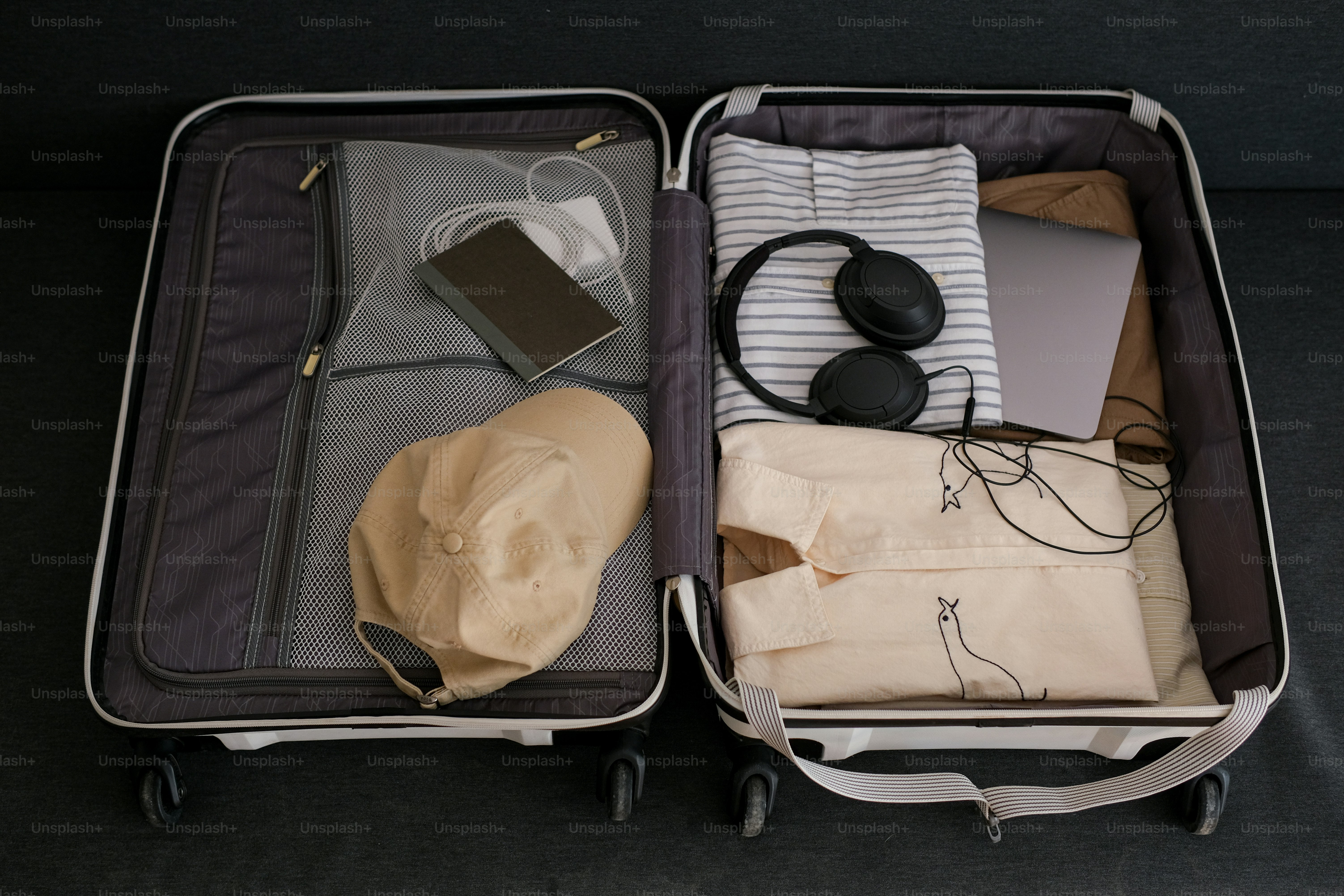 An open suitcase packed with clothes, electronics, and a cap.