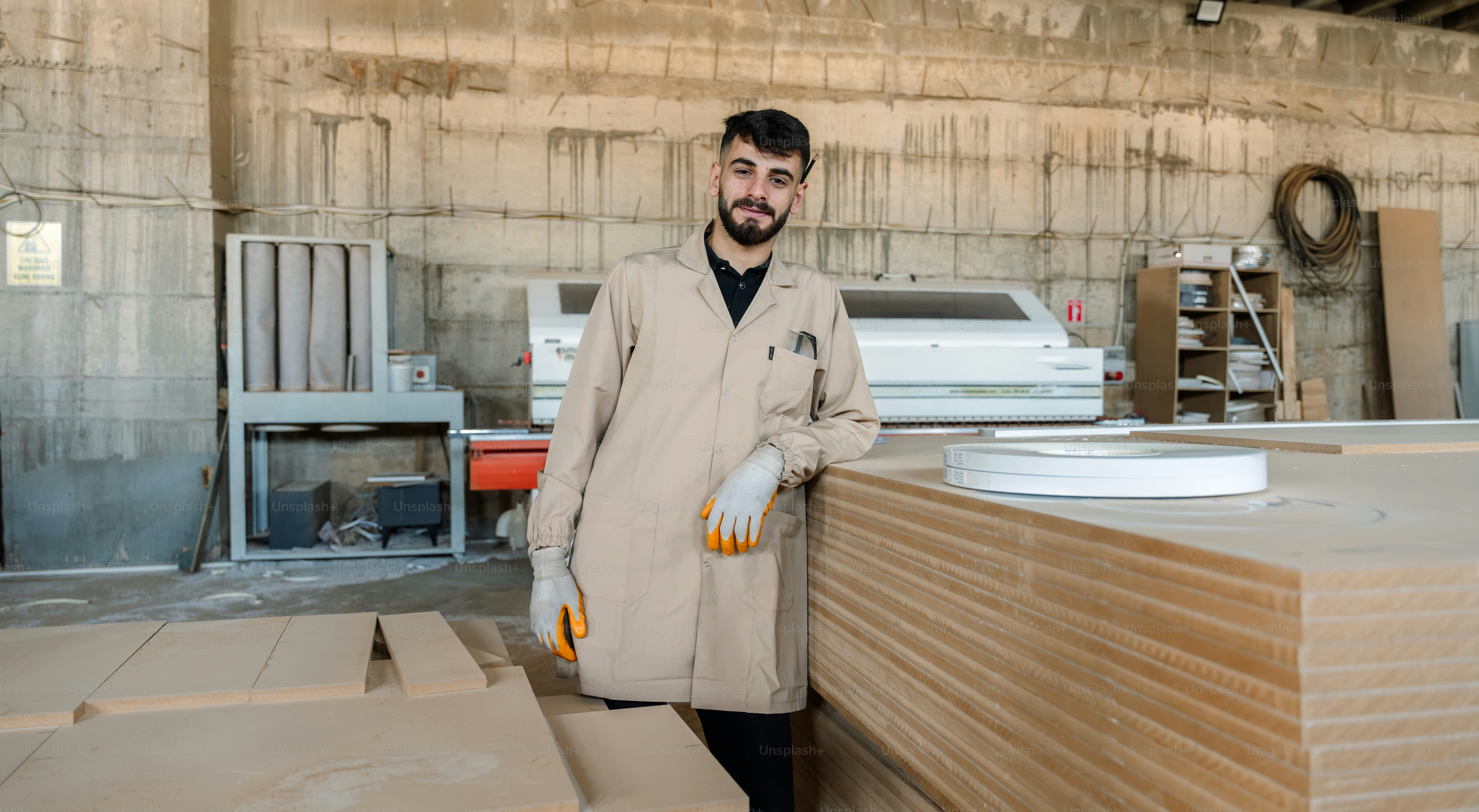 Man in coat and safety glasses leans on wood stack.