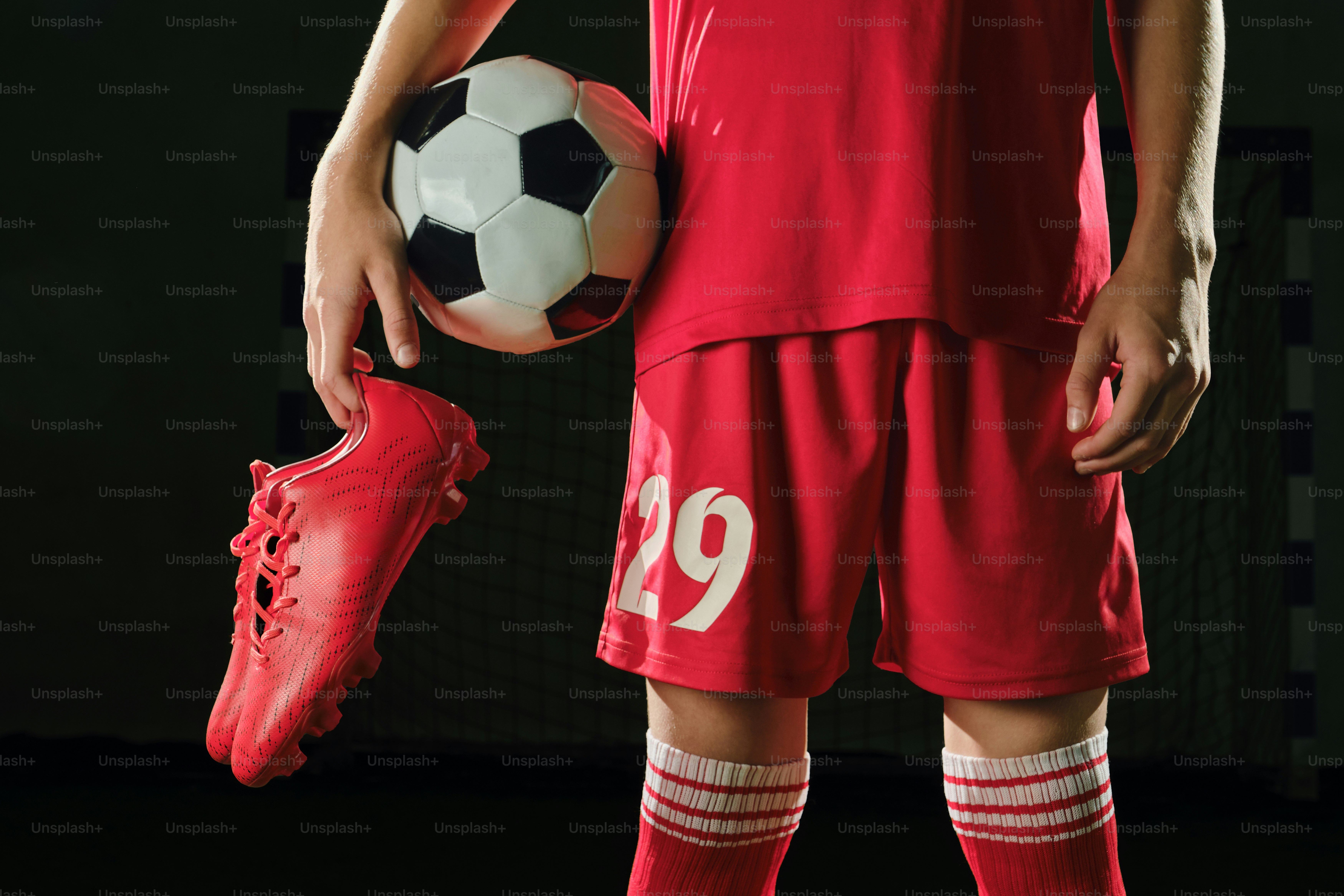 Pemain sepak bola berseragam merah memegang bola dan cleat.