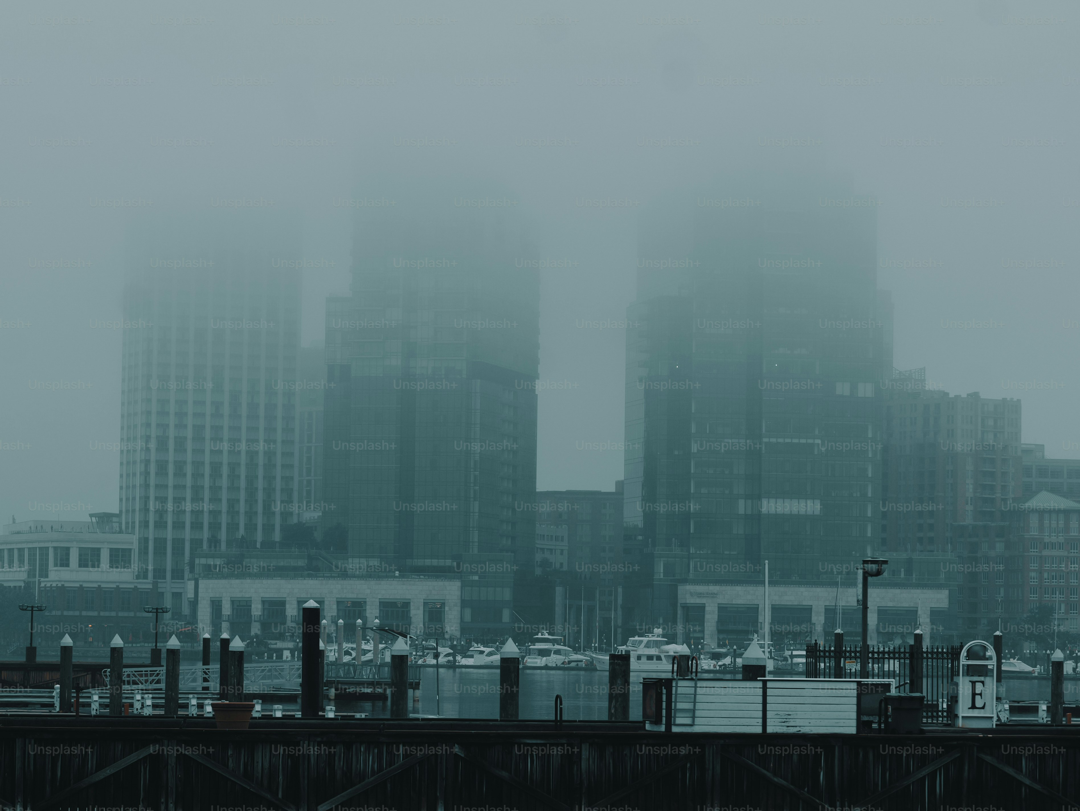 Dense fog obscures a city skyline and waterfront with boats.
