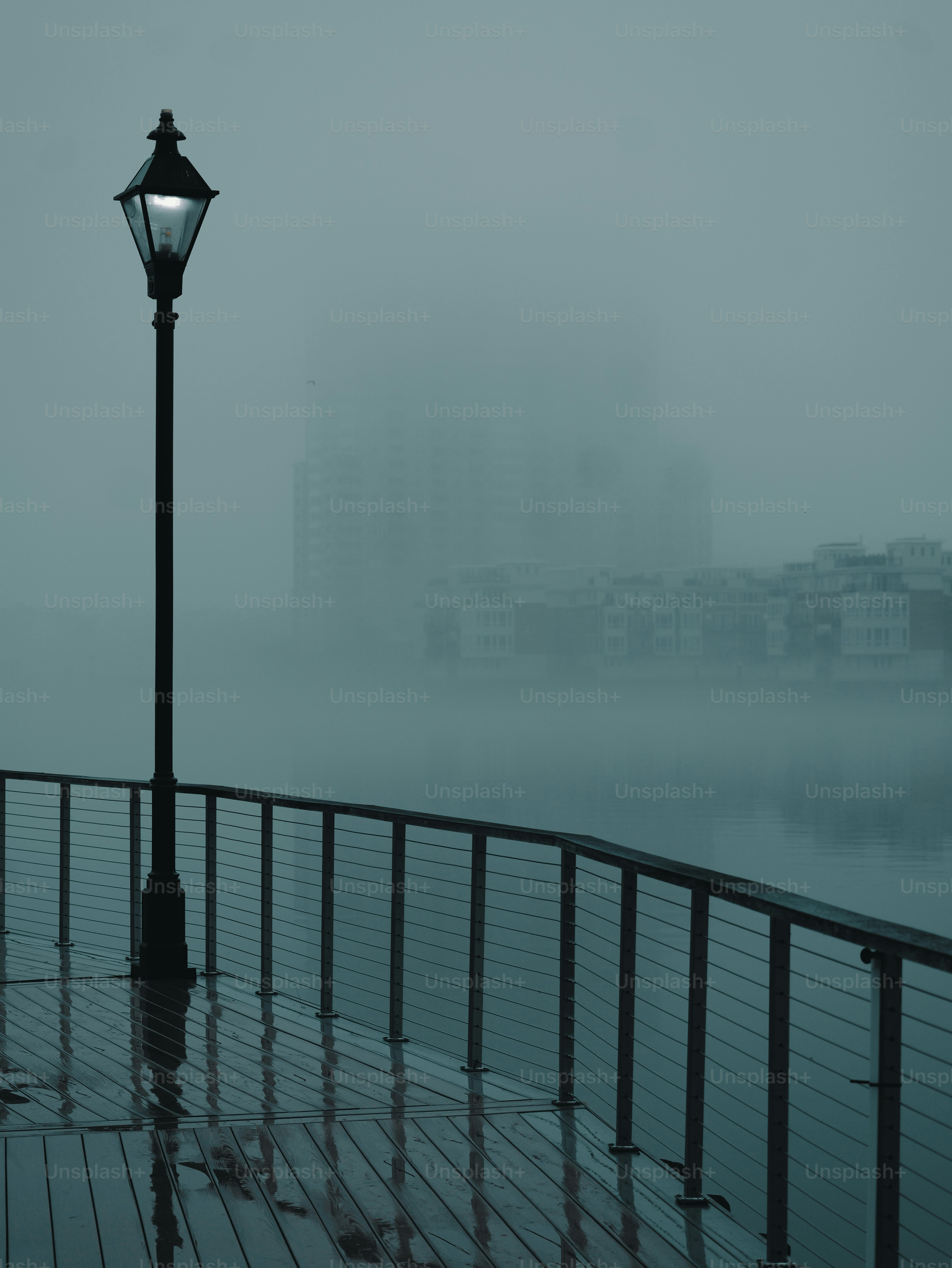 A lit street lamp on a foggy pier overlooking water.