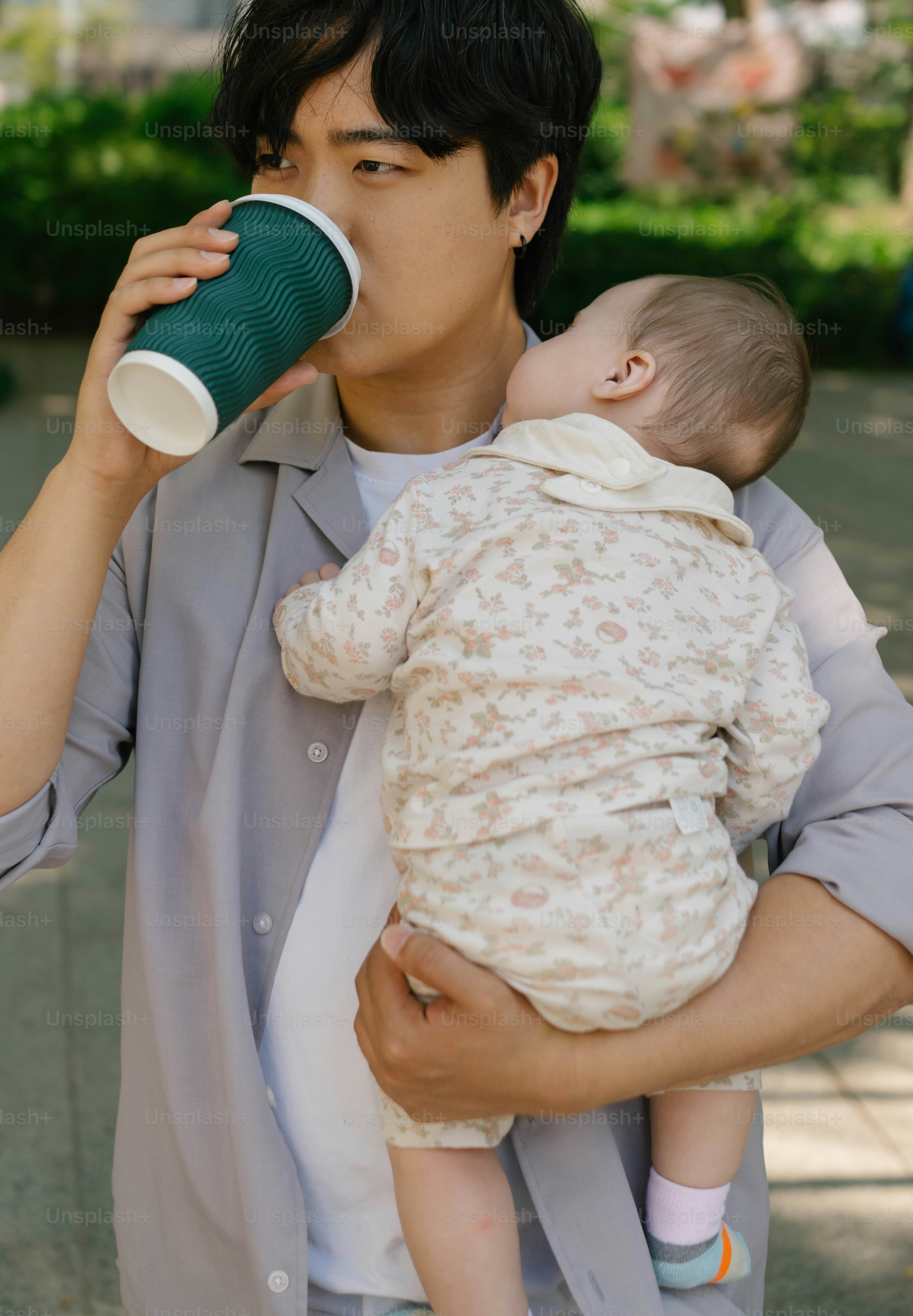 Ayah minum dari cangkir sambil menggendong bayi yang sedang tidur.