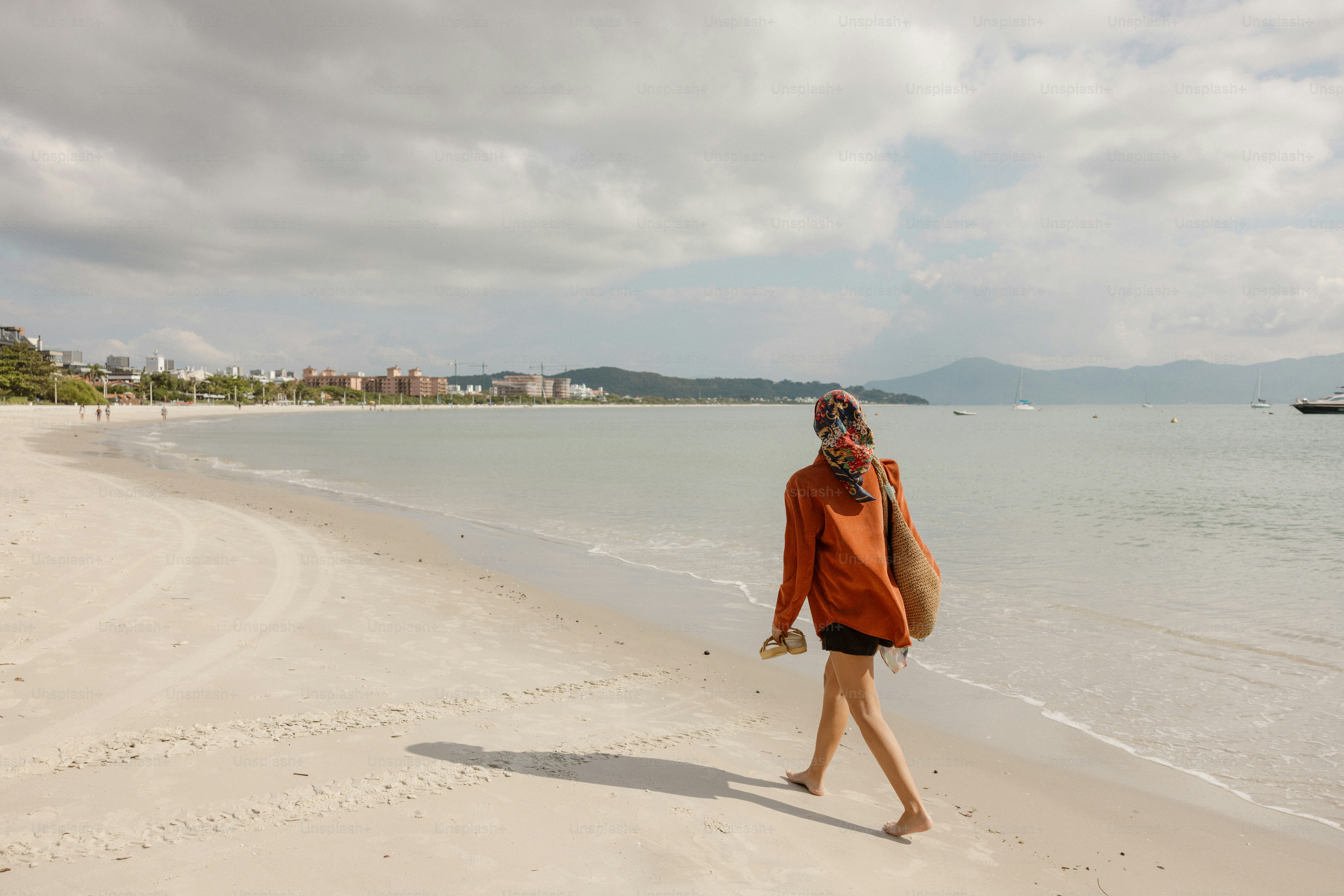 Woman walking barefoot on a calm beach with distant buildings.