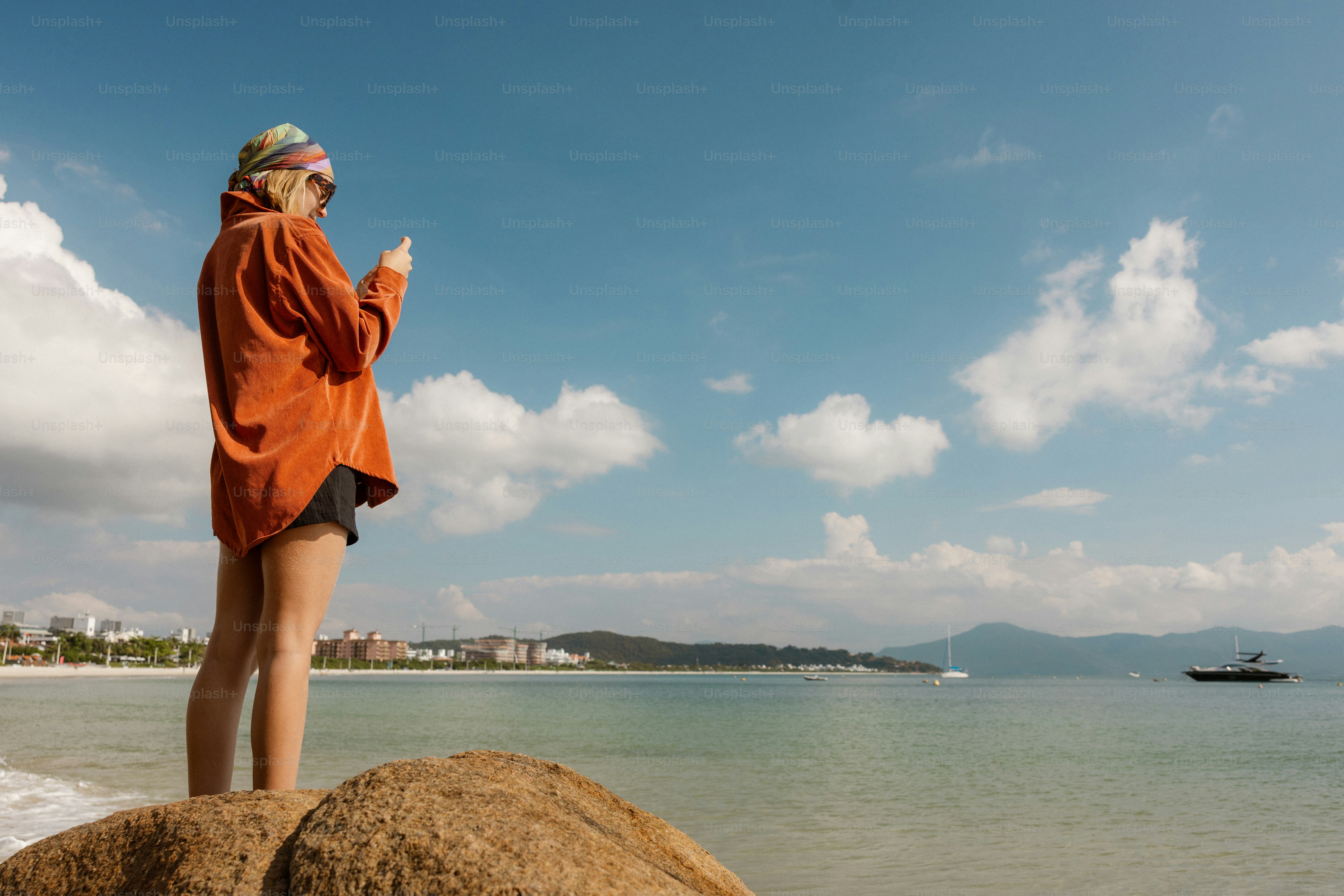 Woman on rock uses phone by the ocean.