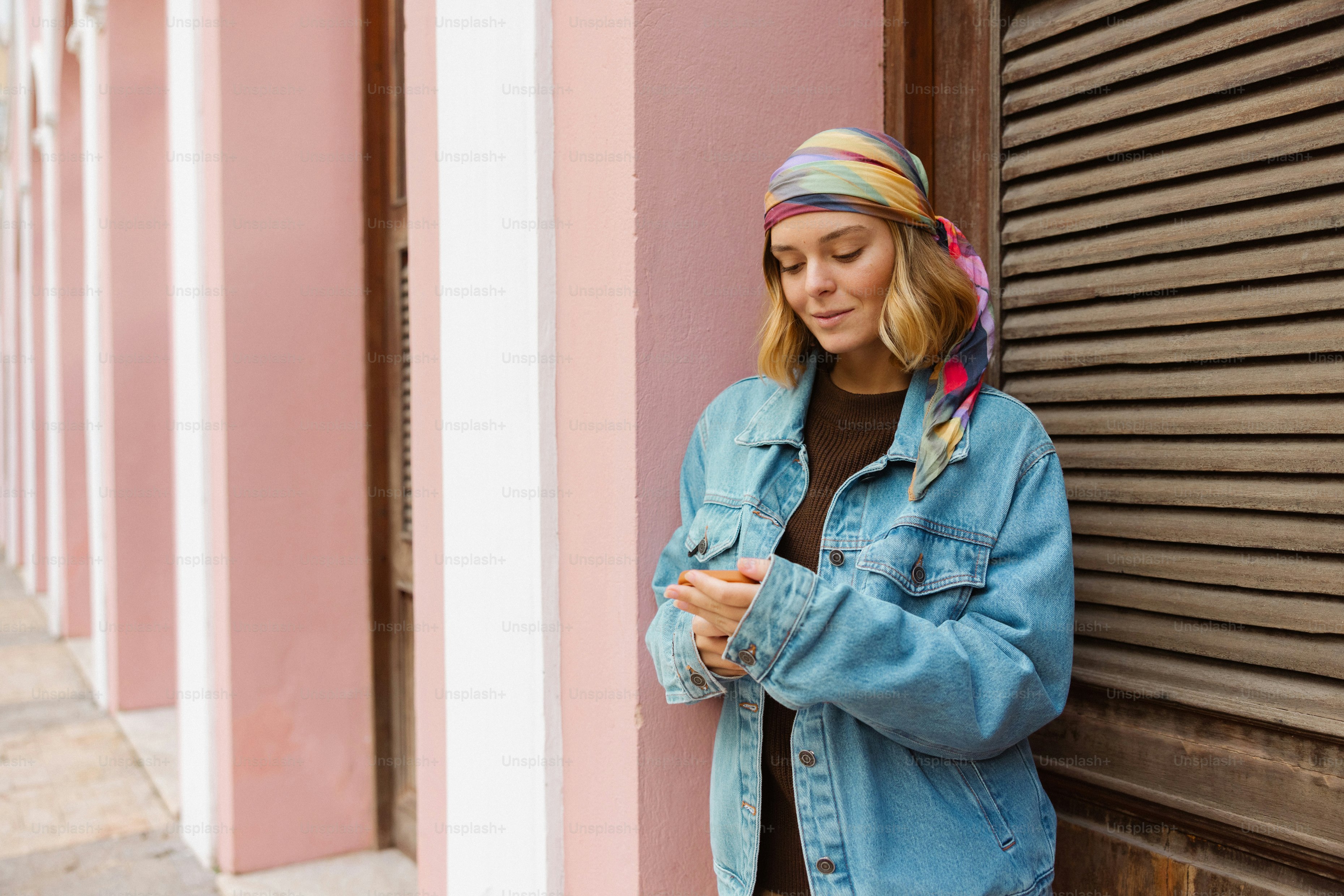 Blonde Frau mit Kopftuch und Jeansjacke an der rosa Wand.