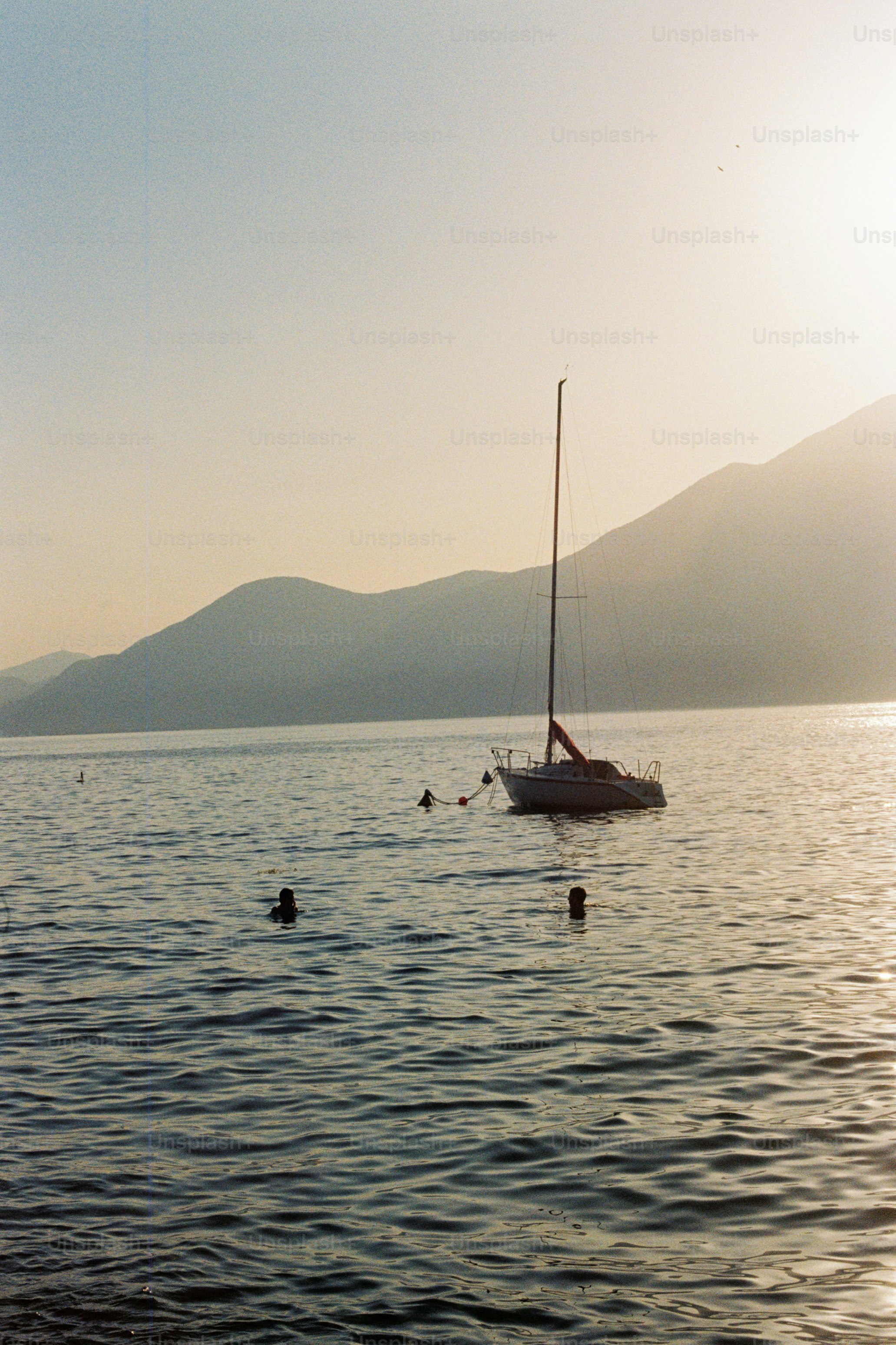 Sailboat anchored in water with mountains and swimmers at sunset.
