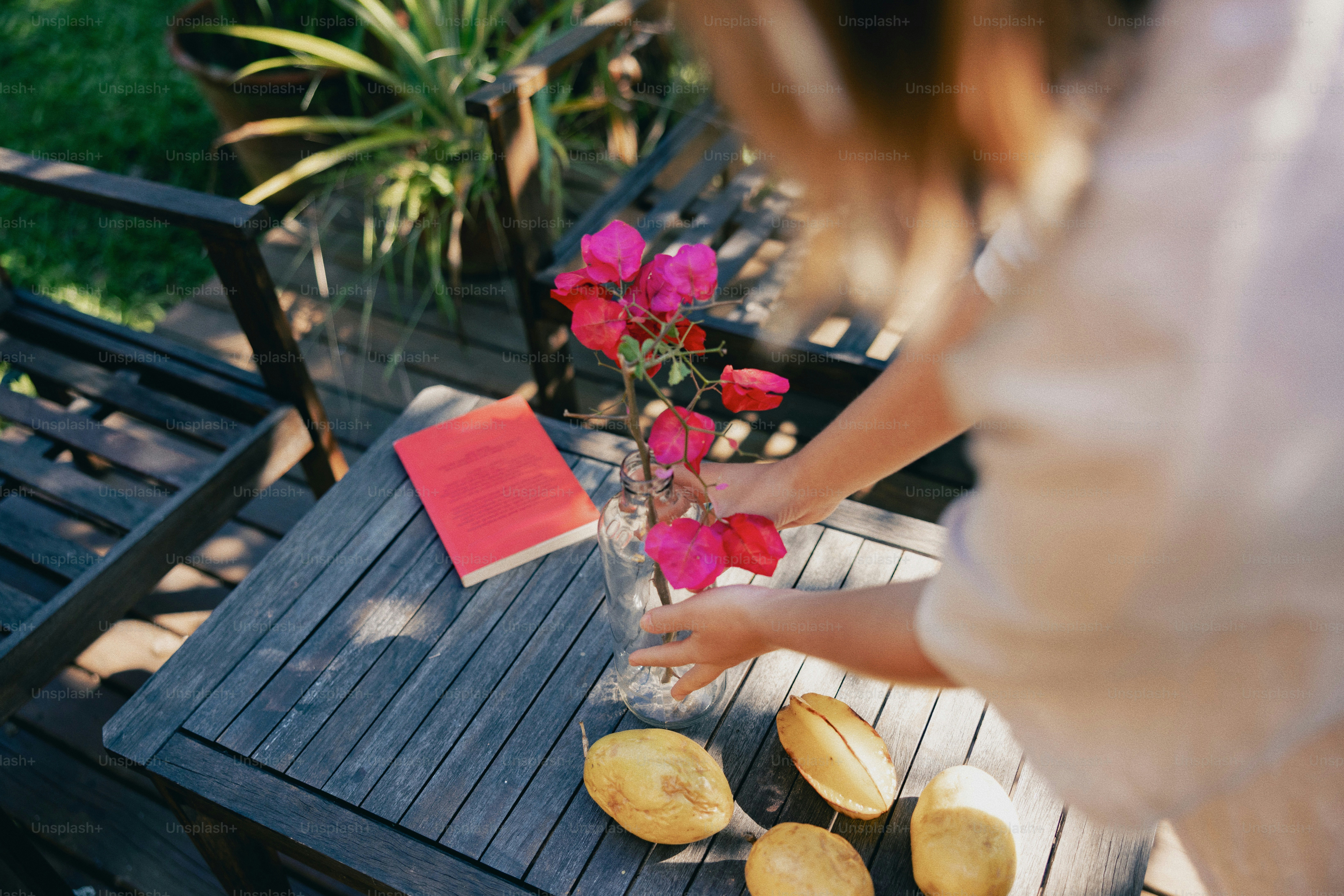 Hands arranging pink flowers, book, and fruit on wooden table.