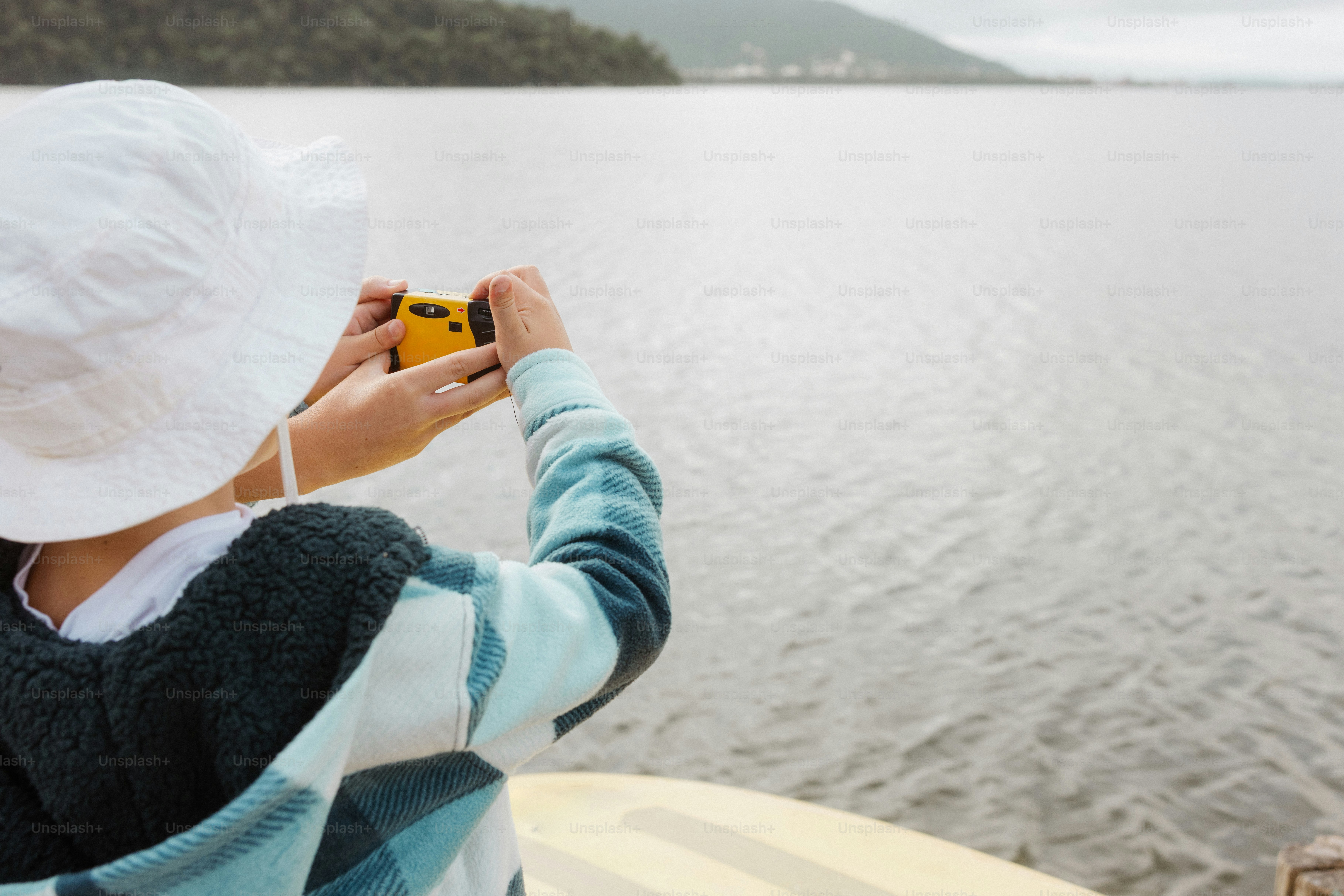 Orang bertopi memotret danau dengan kamera kuning.