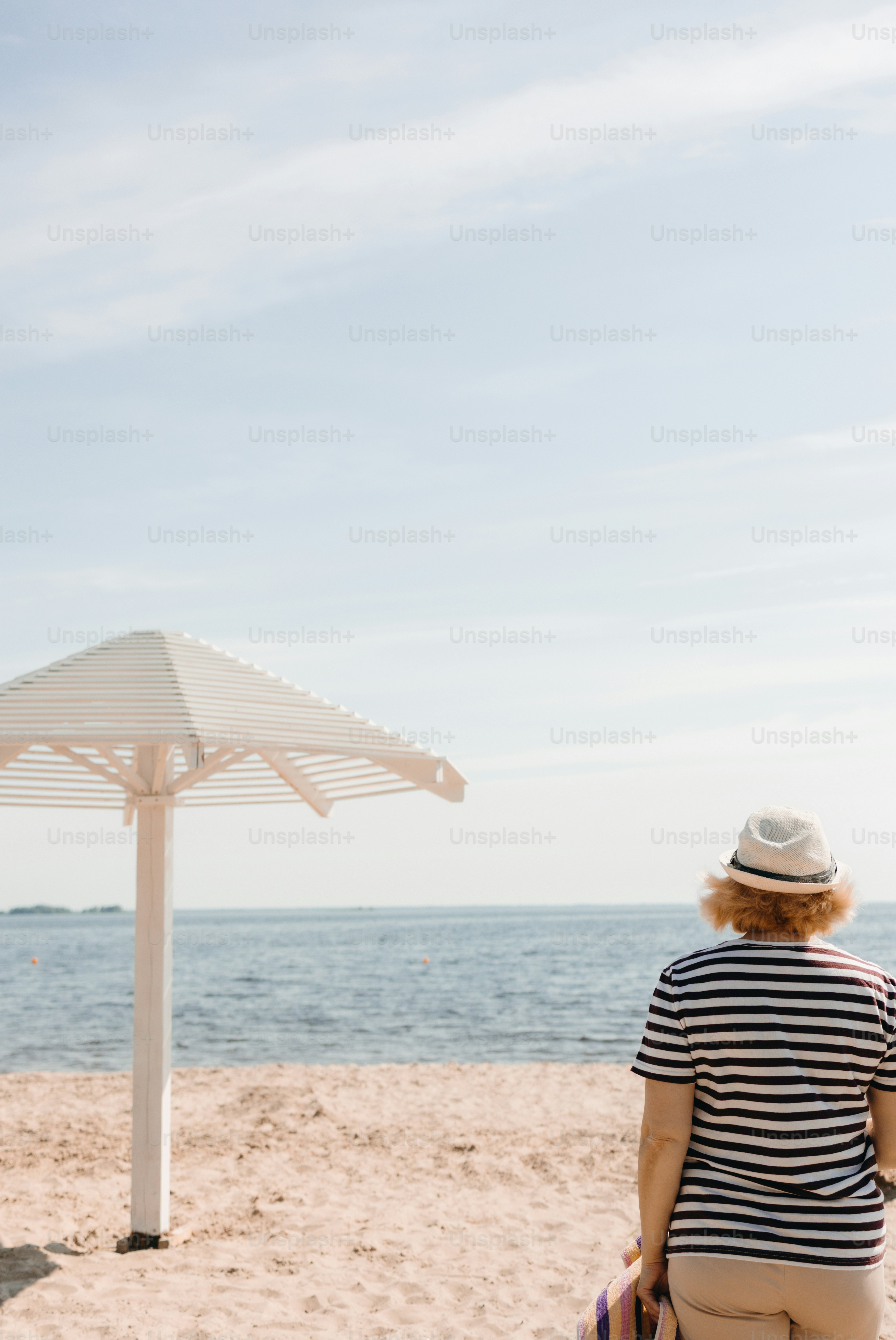 Woman in hat looks at ocean from sandy beach.