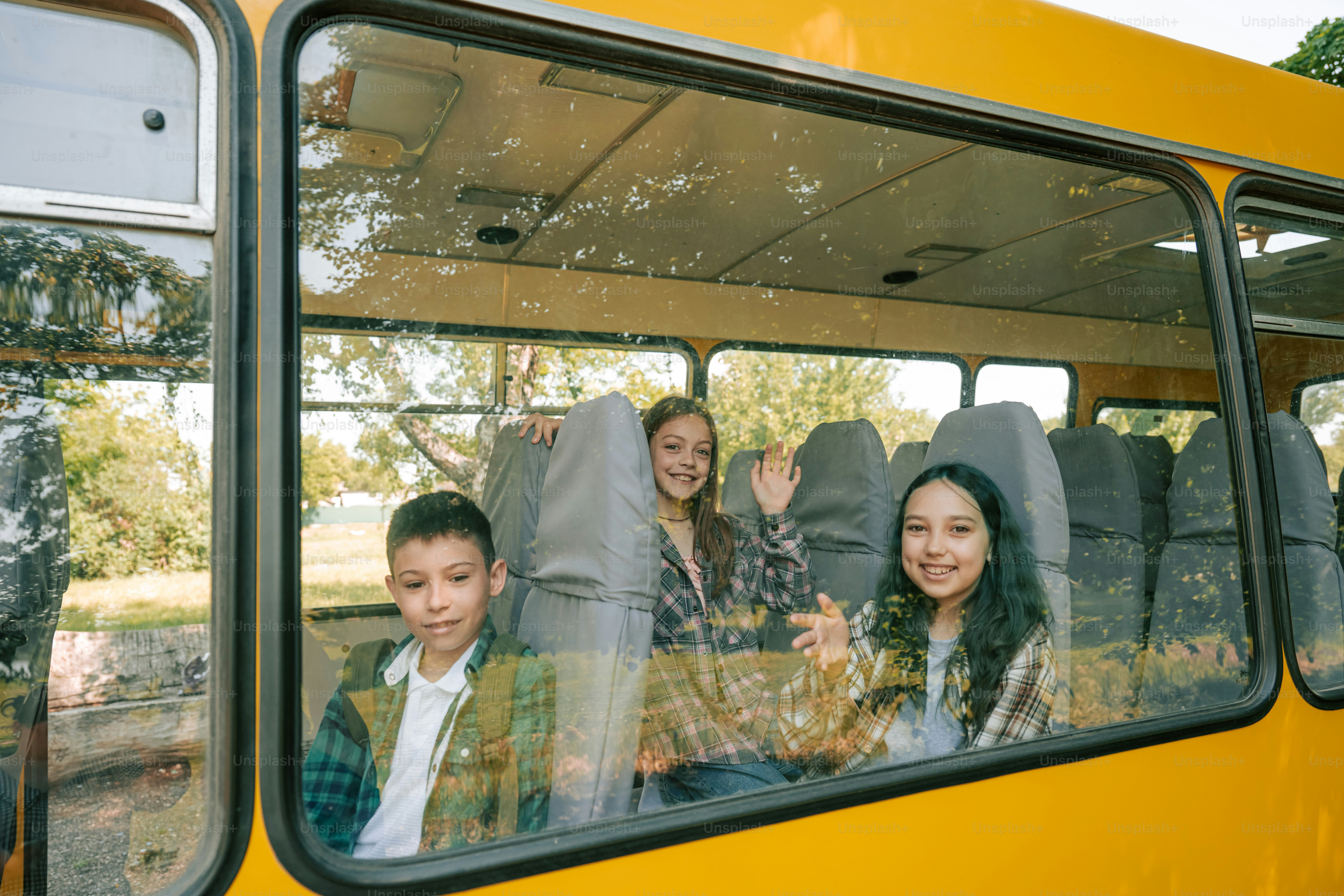 Three smiling children looking out a yellow bus window