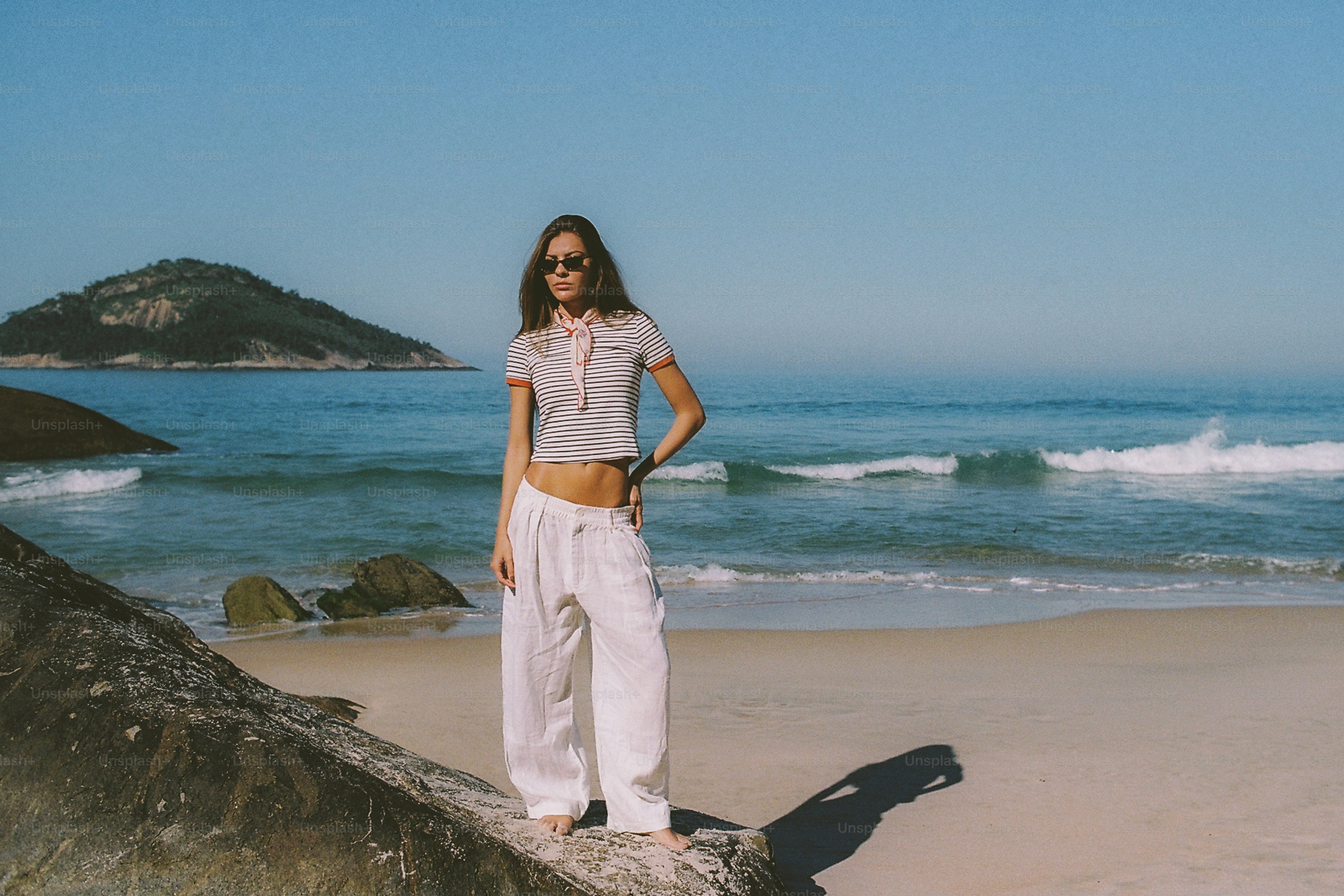 A woman stands on a rock at a sunny beach.