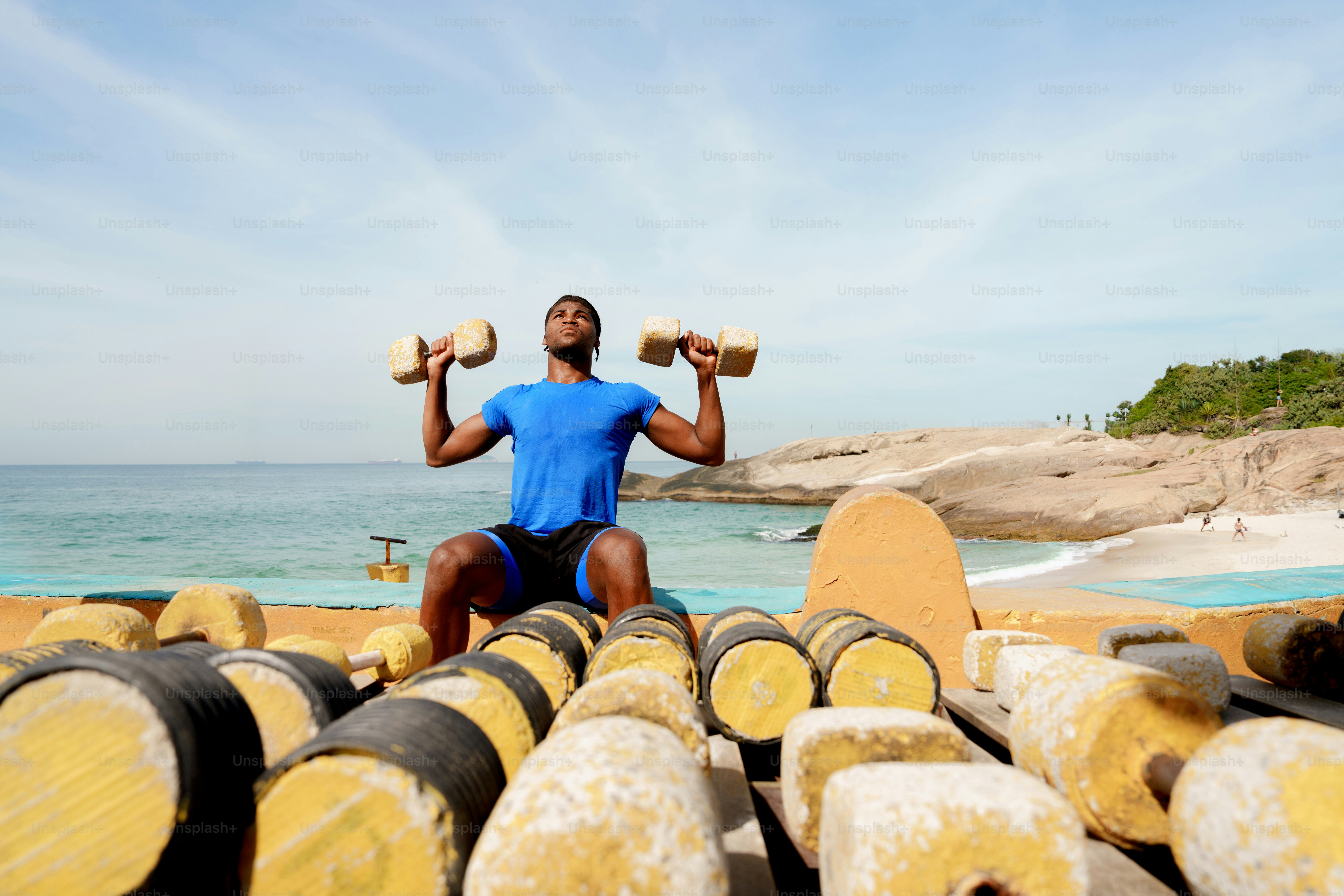 A man lifts wooden dumbbells on a beach with logs.