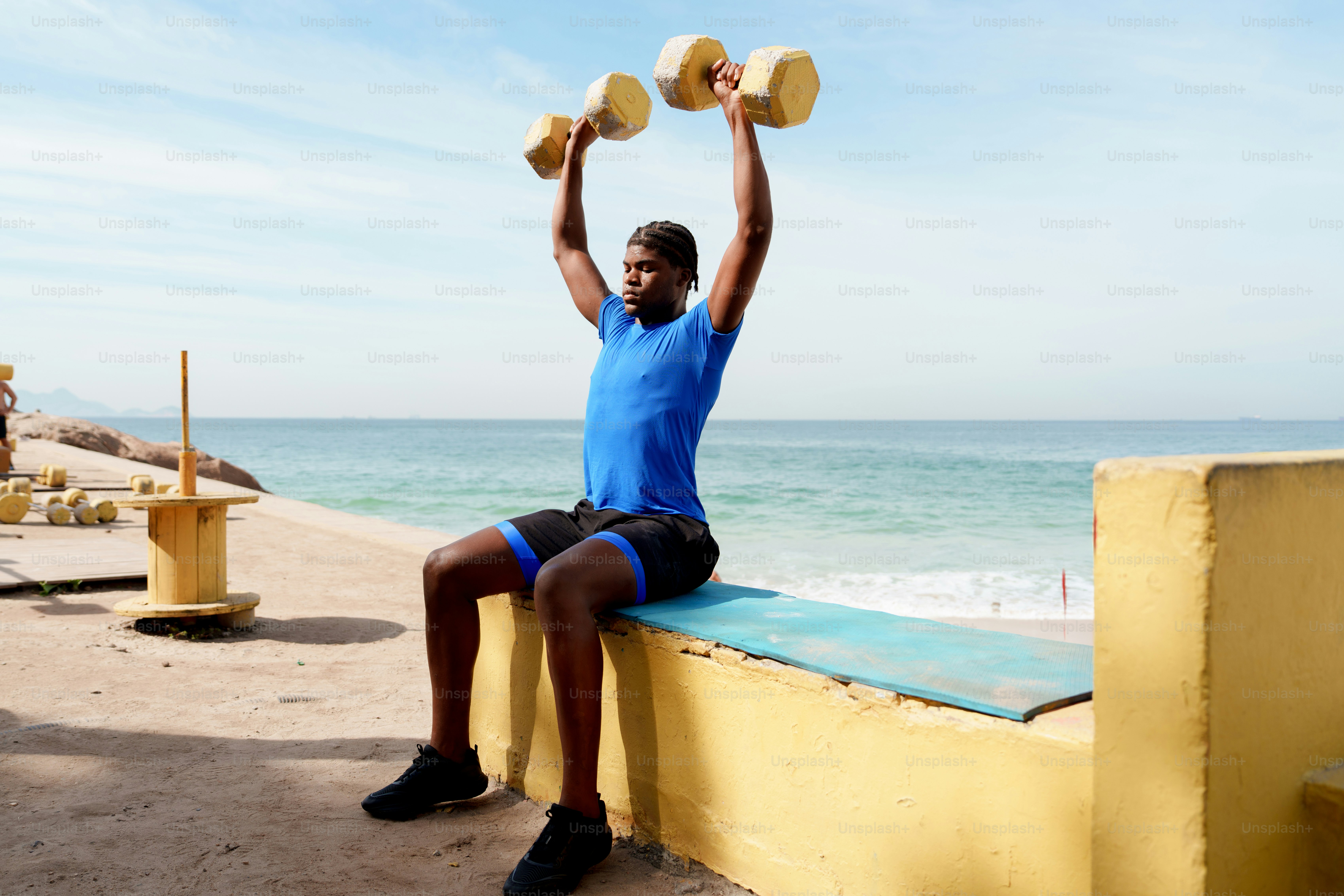 Man doing shoulder press with dumbbells outdoors by the sea.