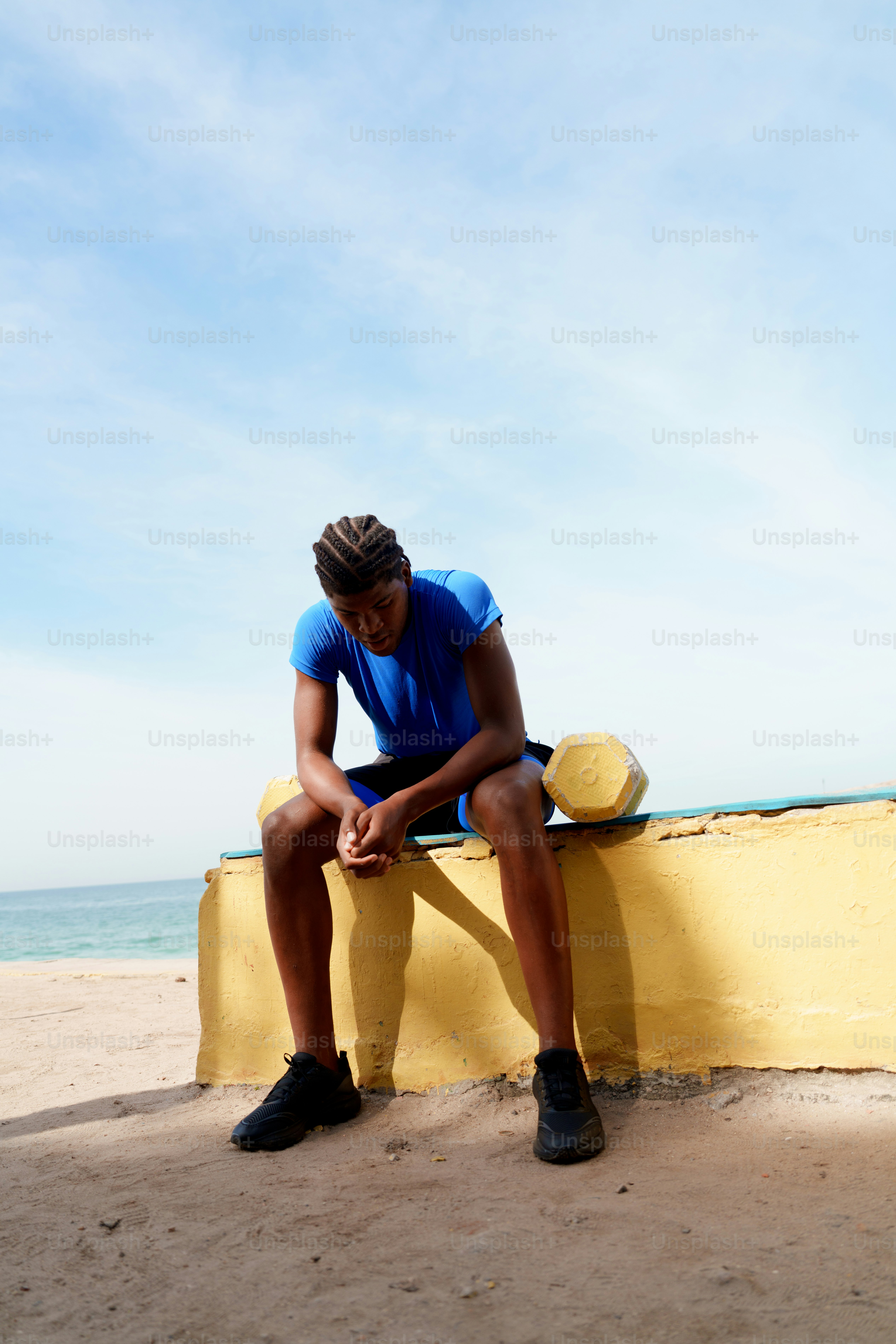 Young person in blue sits on yellow wall by ocean.