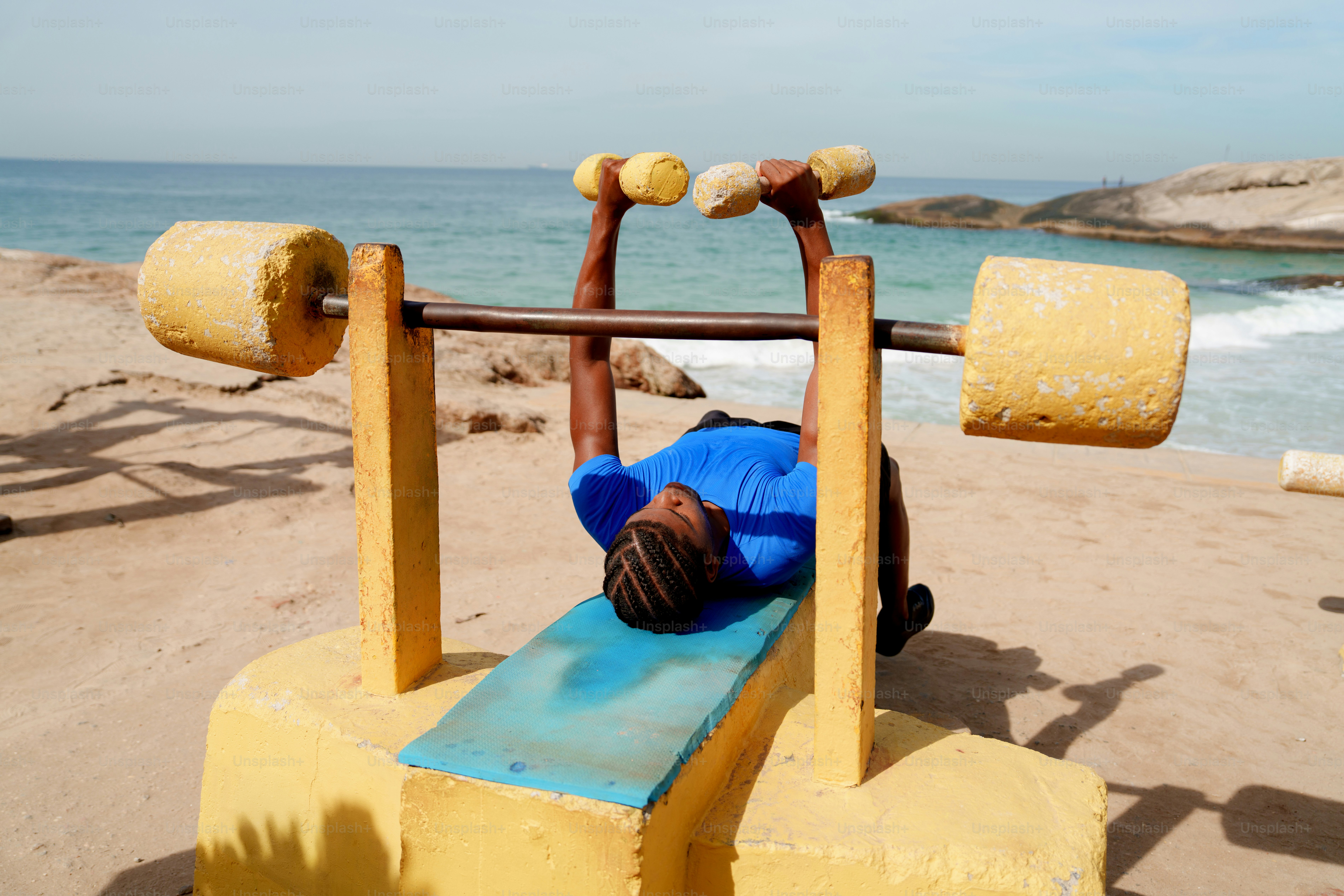 A man bench presses homemade weights on a sunny beach.