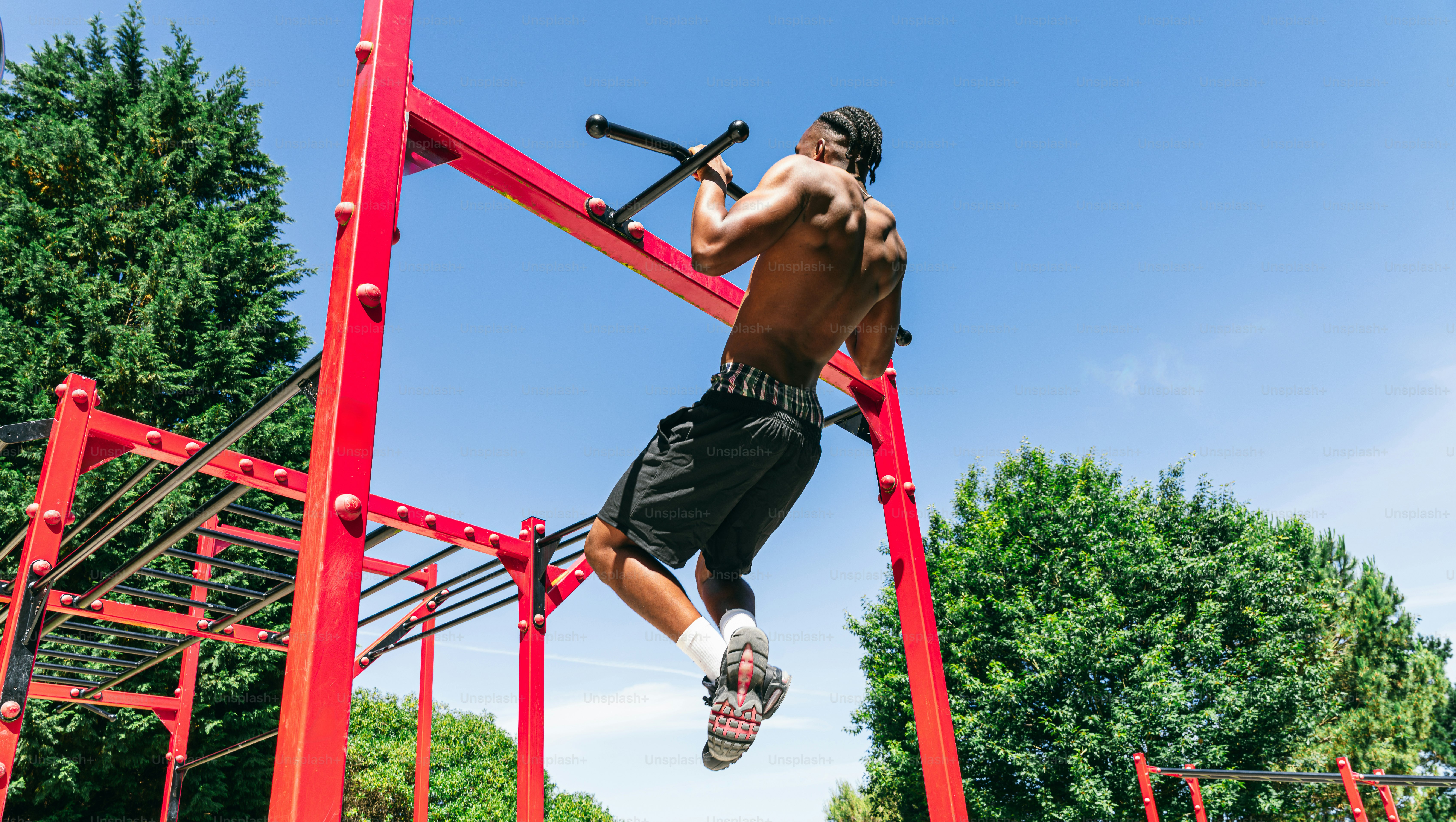 Pria bertelanjang dada melakukan pull-up pada peralatan olahraga luar ruangan merah.
