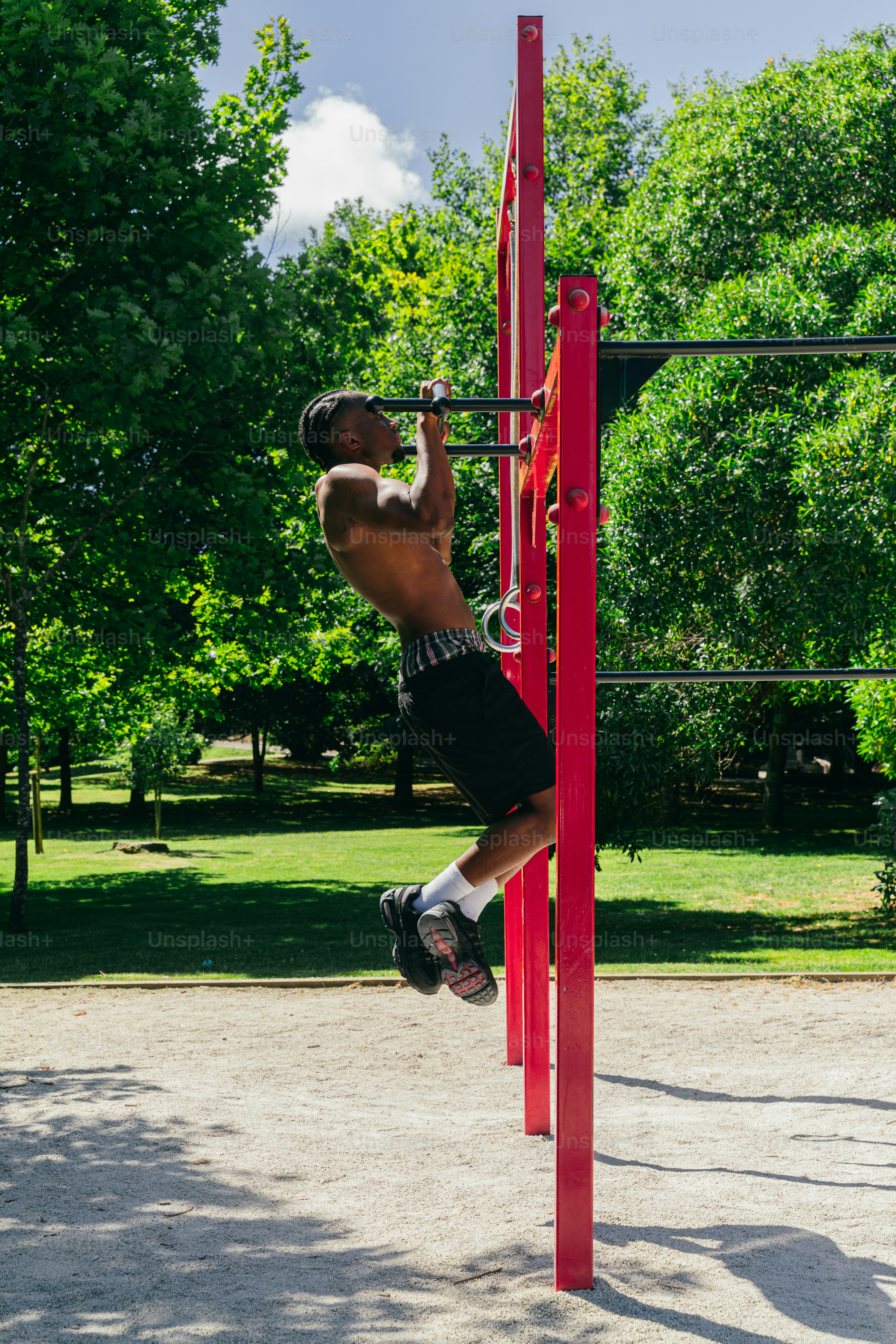 A shirtless man doing pull-ups on an outdoor red bar.