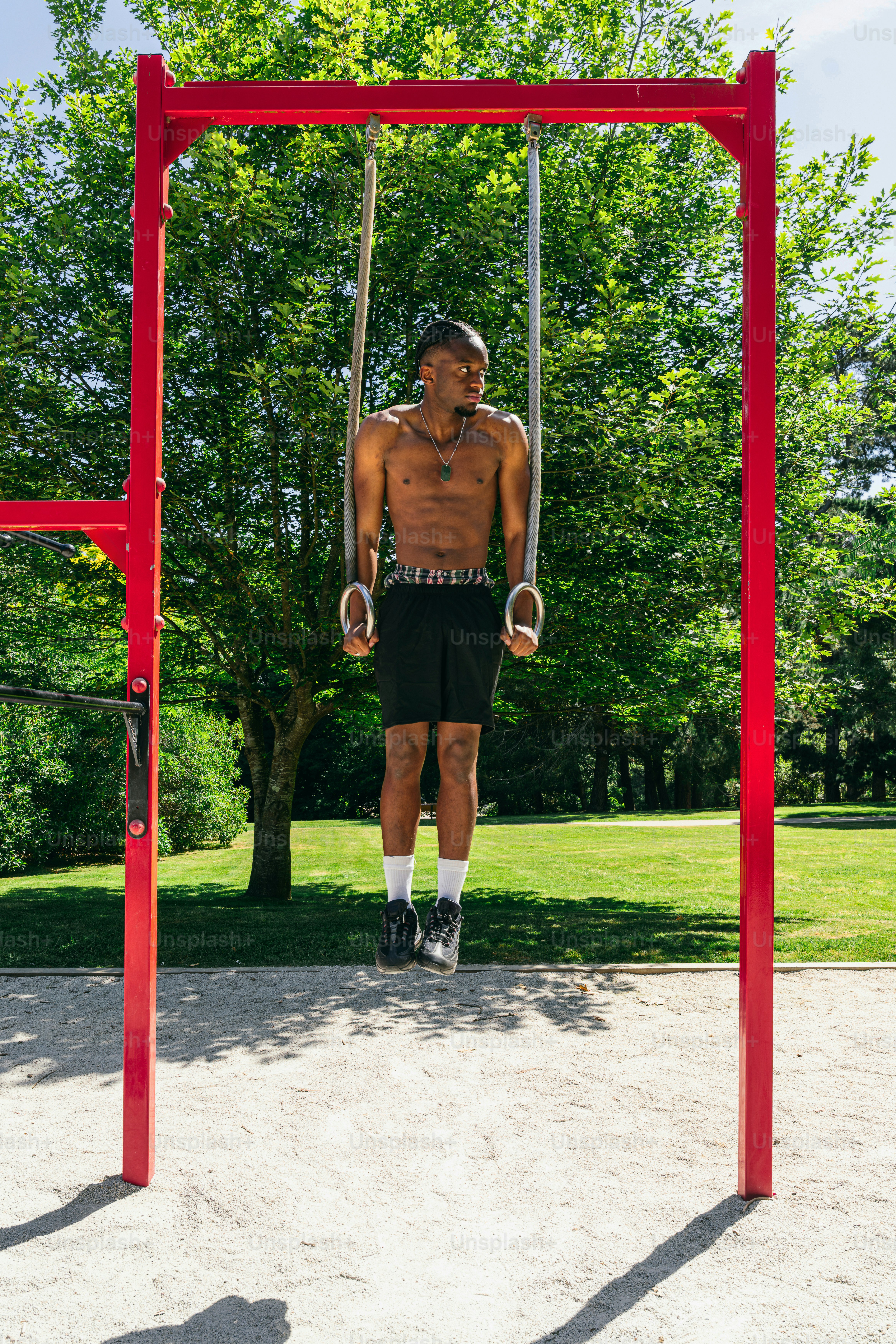 Shirtless man exercising on gymnastic rings outdoors.
