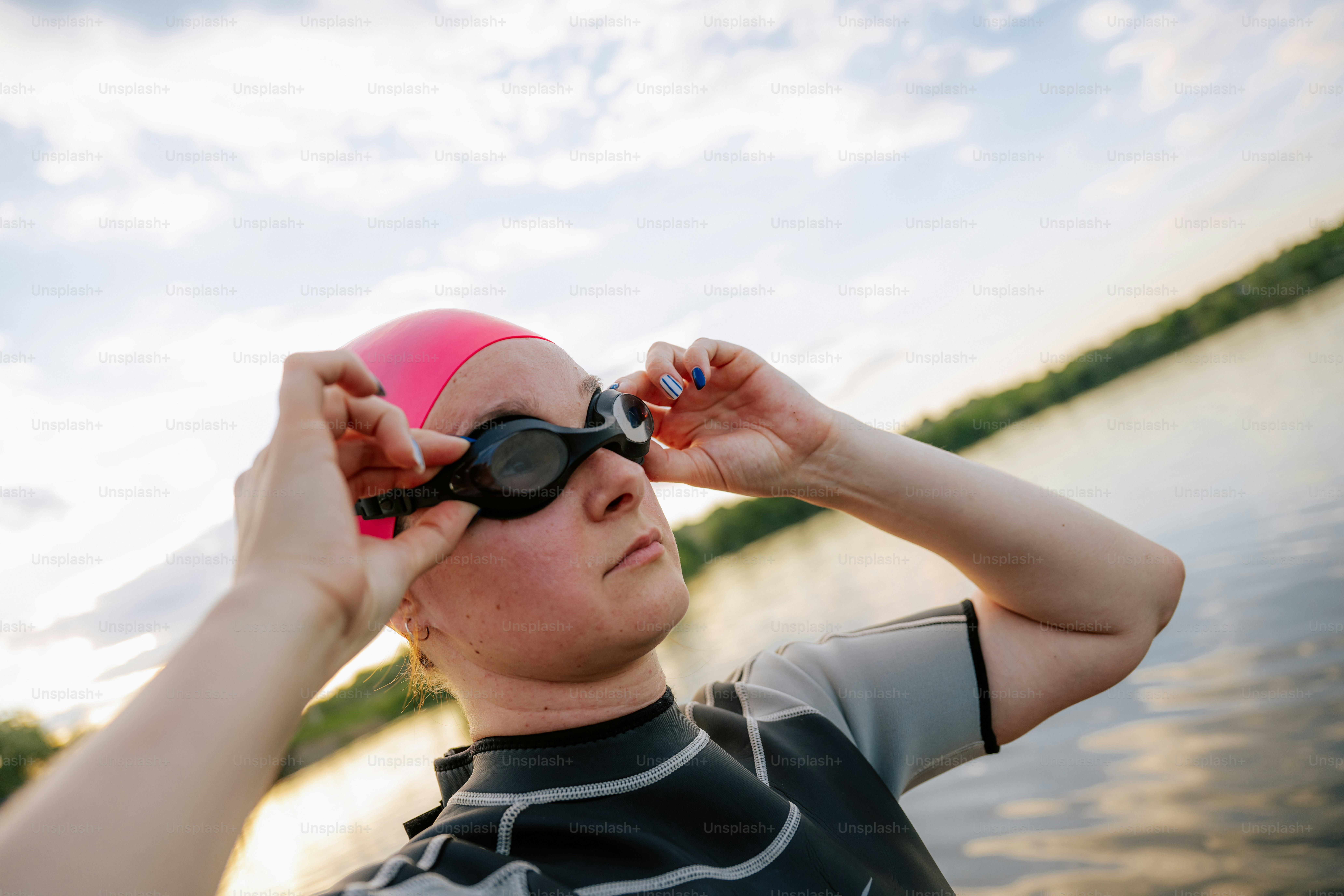 Femme en combinaison et casquette rose ajustant des lunettes près de l’eau.