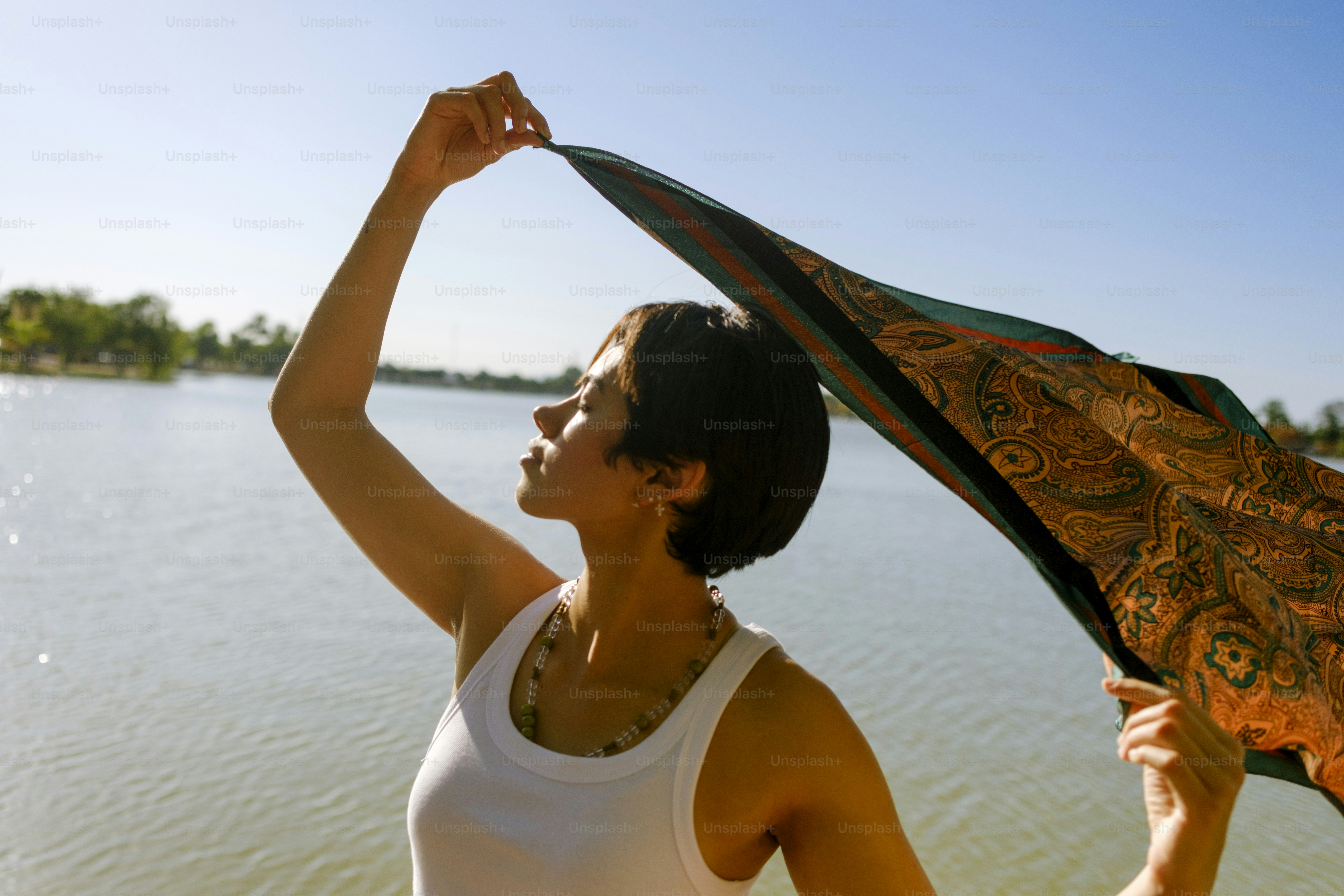 Woman holding patterned scarf by a sunny lake.
