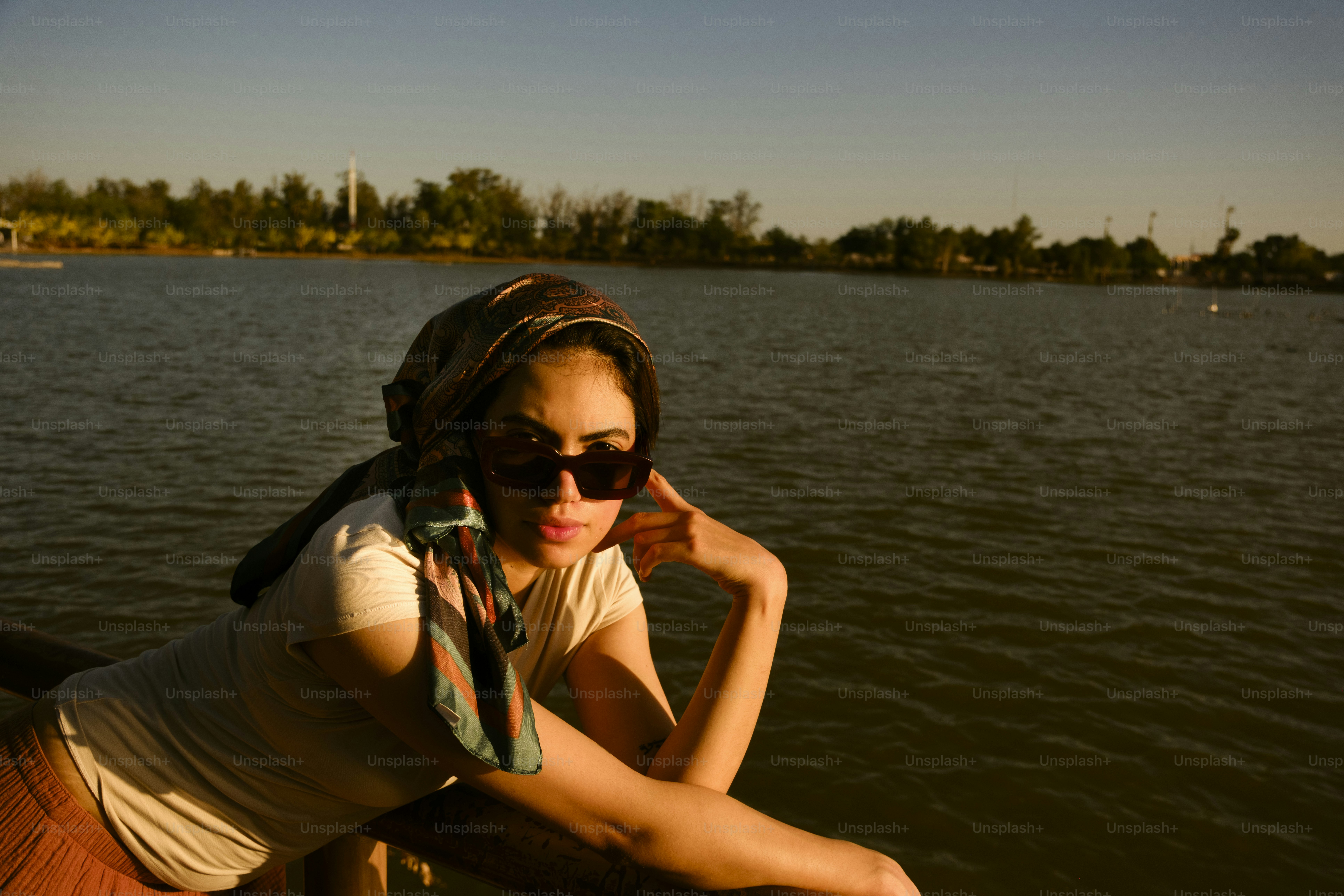 Woman in sunglasses and scarf by a sunlit lake.