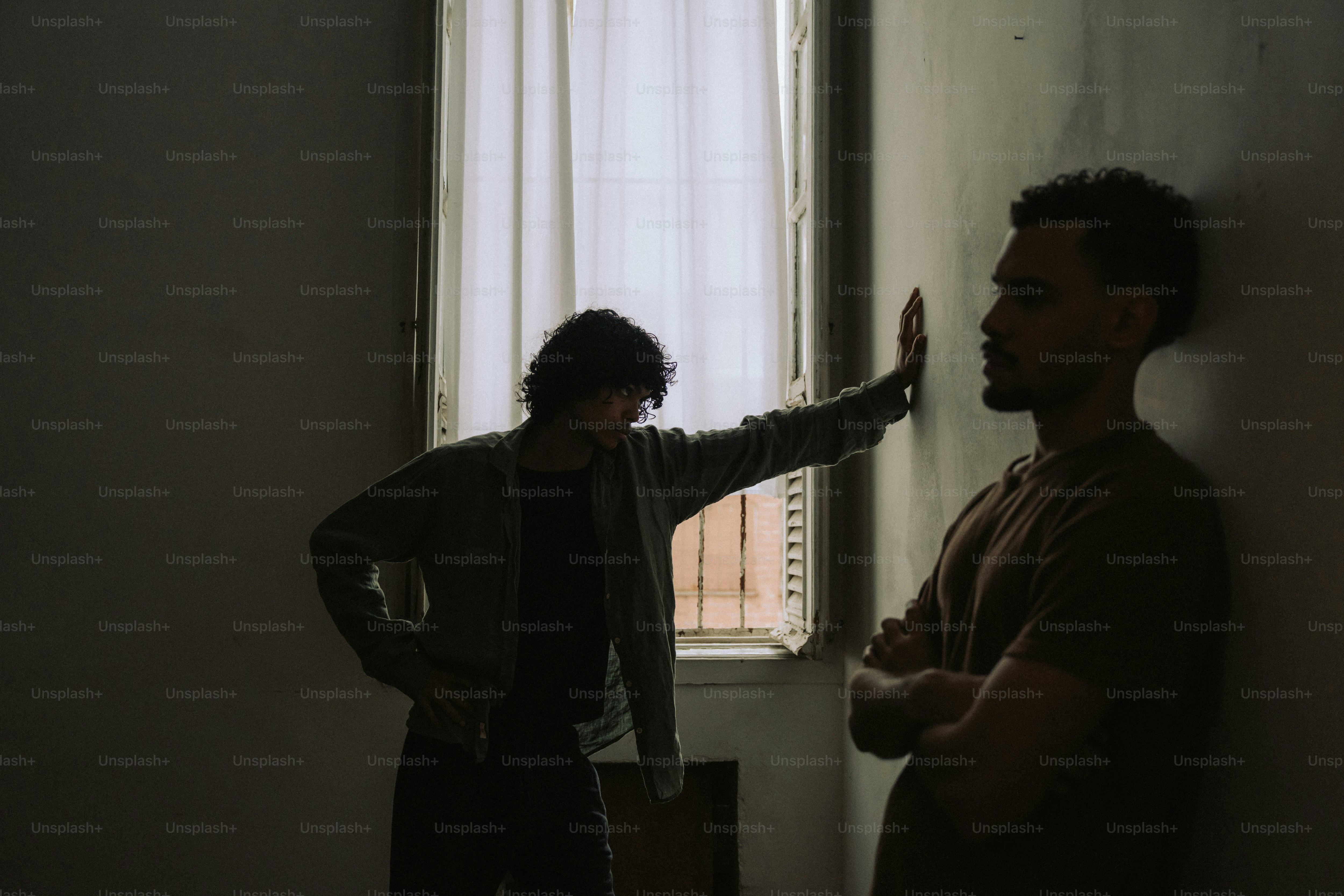 Two men in dark room, one's arm against wall.