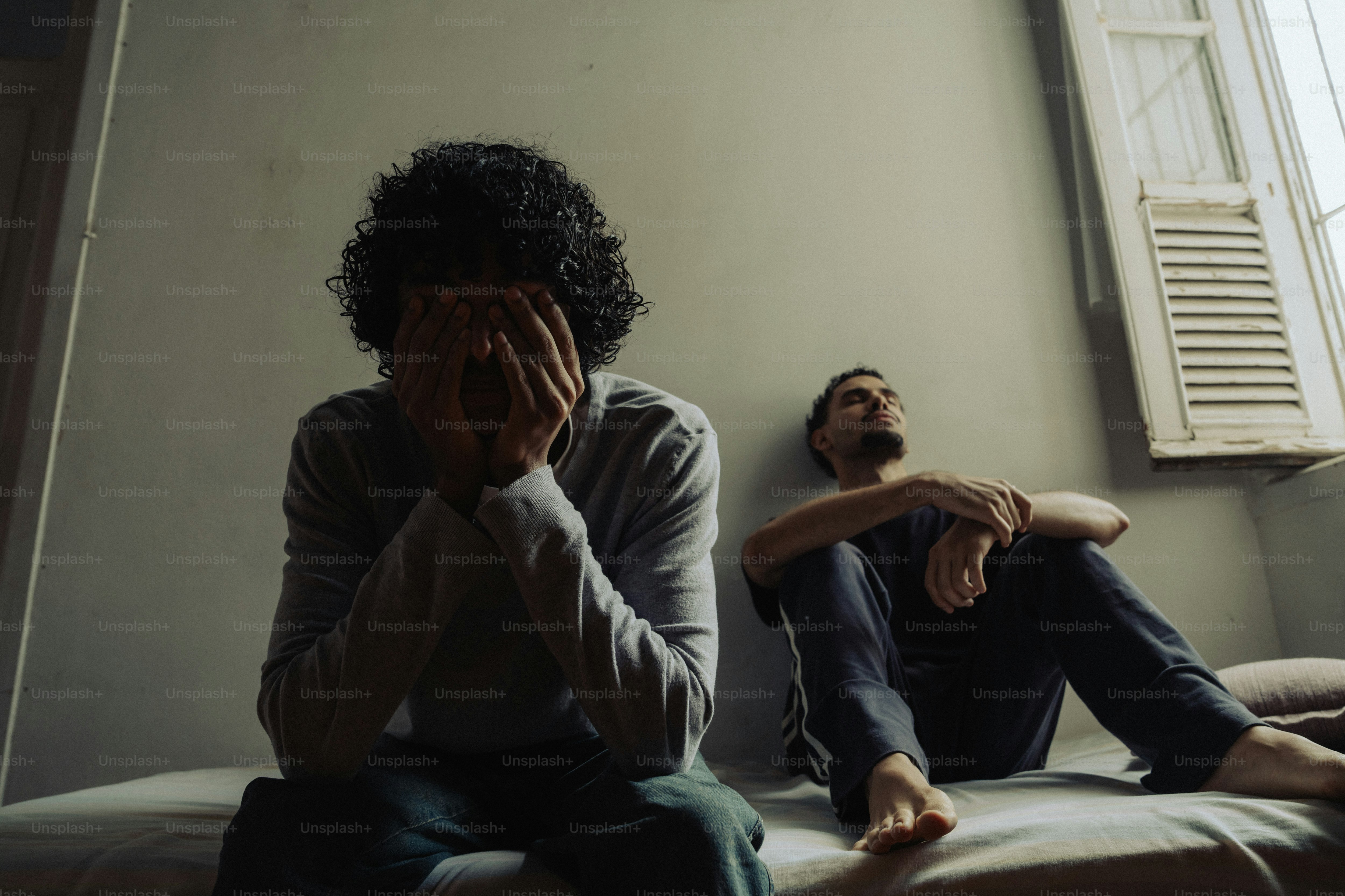 Man covering face in distress, another sits pensively