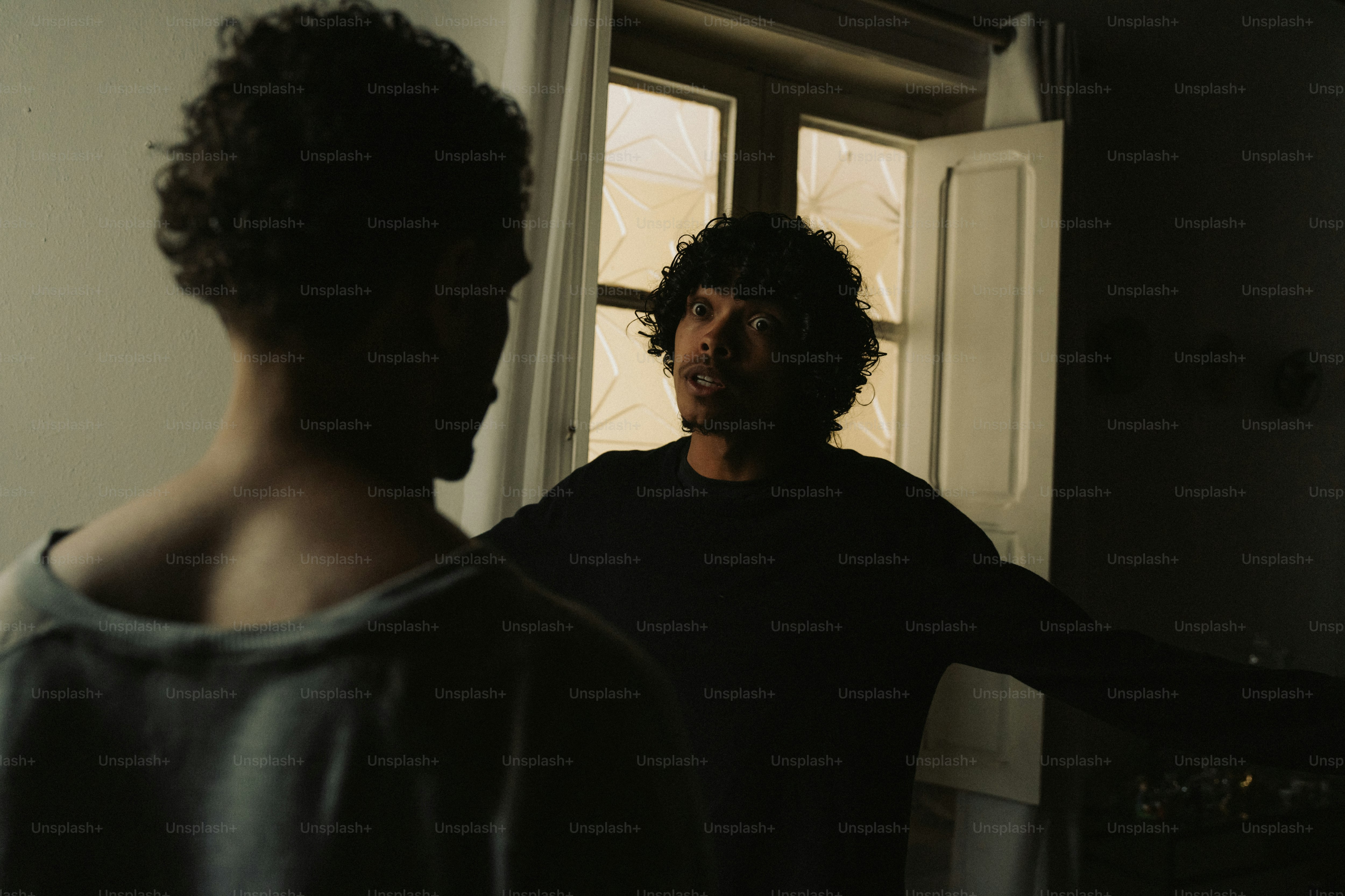 Two men talking indoors near a window.