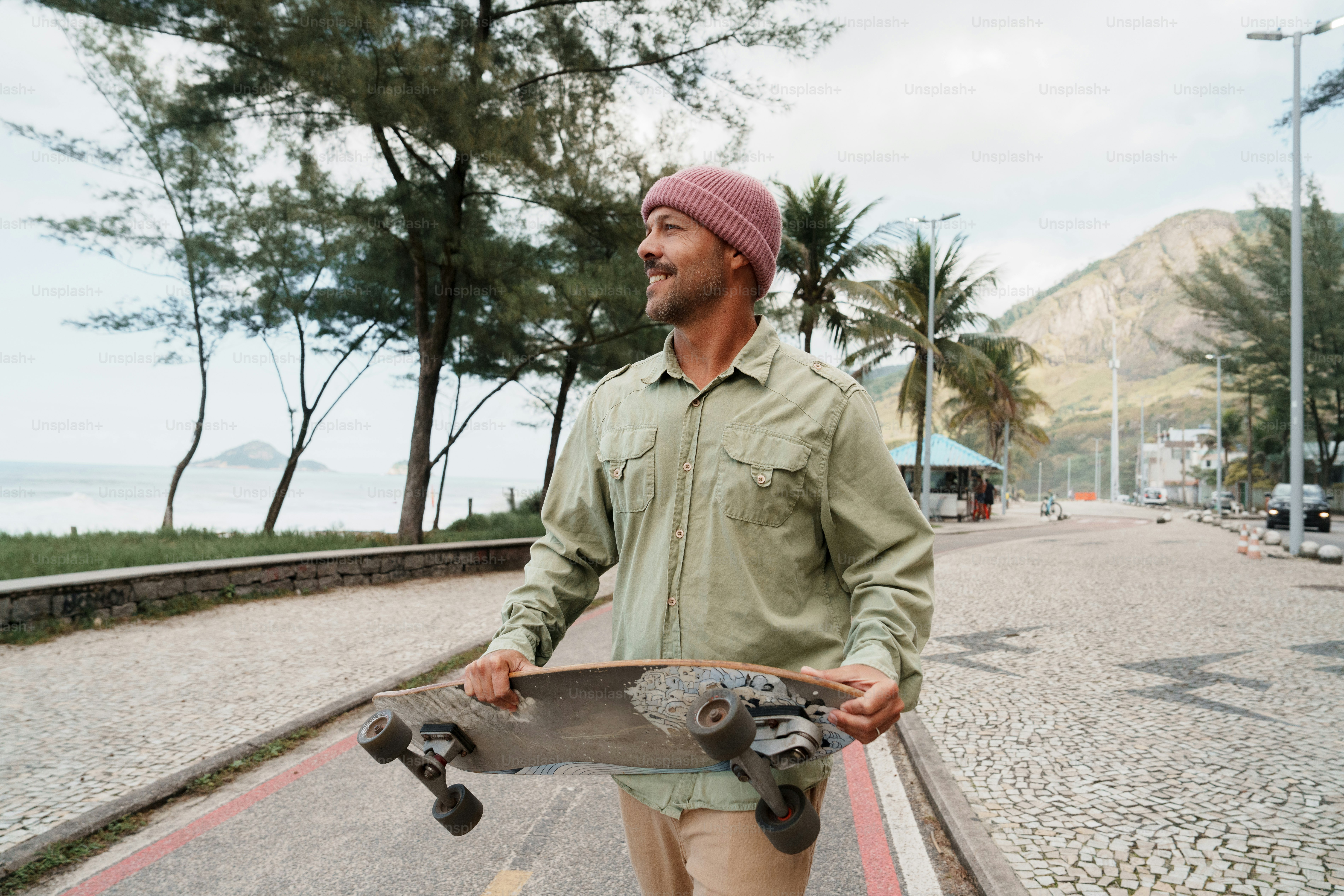 Man in beanie holding skateboard on beachside path