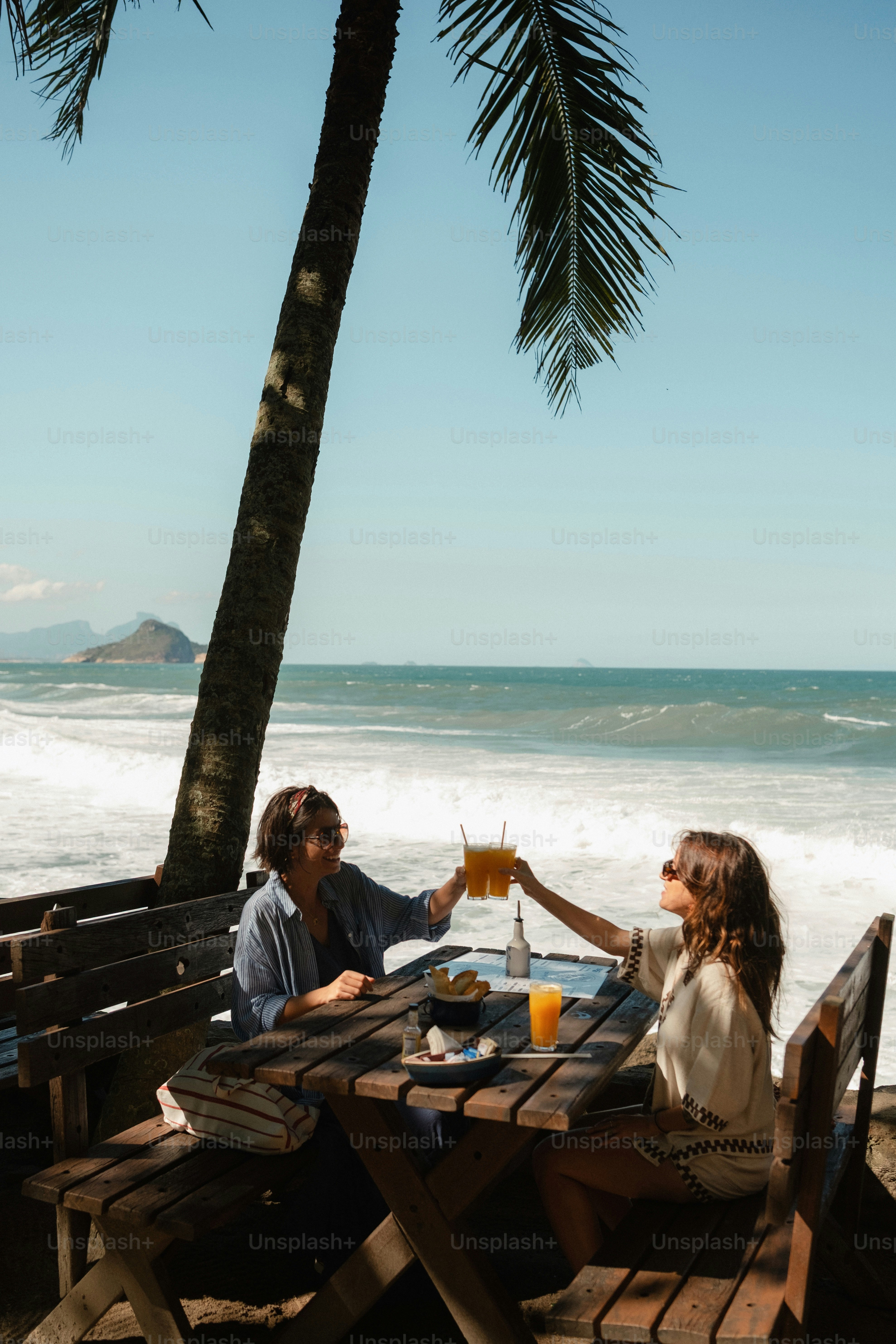 Women toasting drinks at beachside table with palm tree