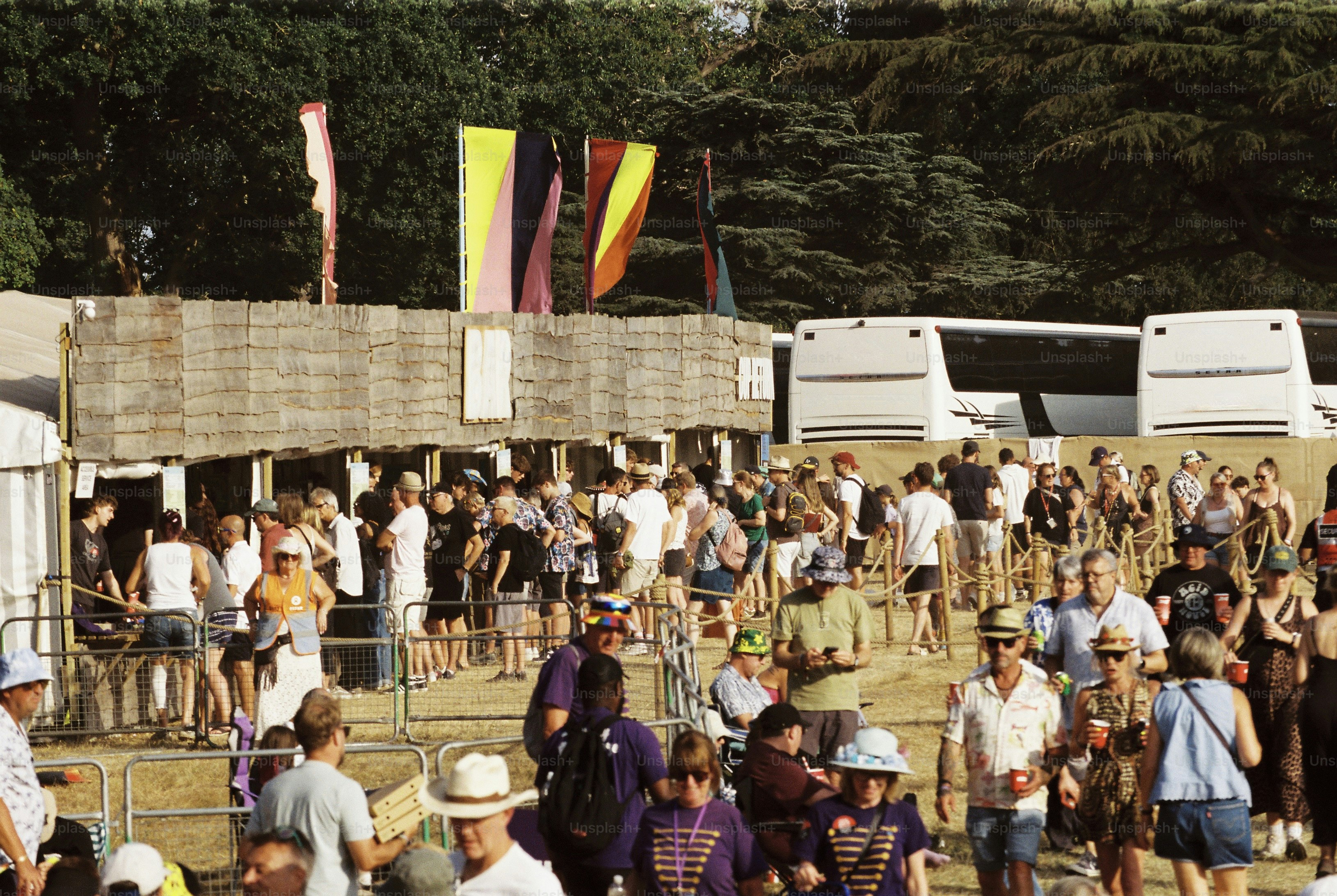 Festivalgoers queue at timber bar under flags, coaches behind