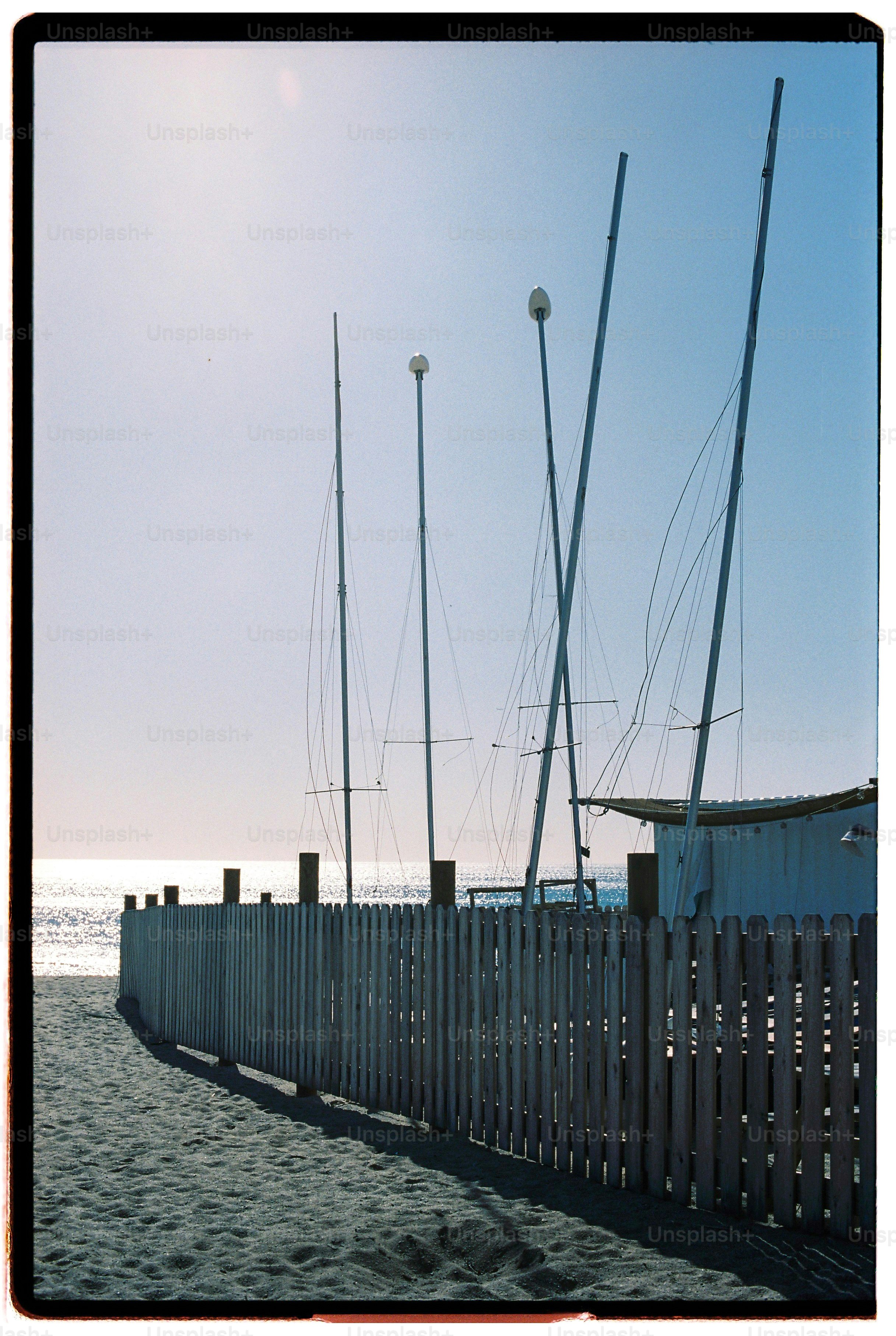 Tall sailboat masts rising behind a white picket fence on a sandy beach.