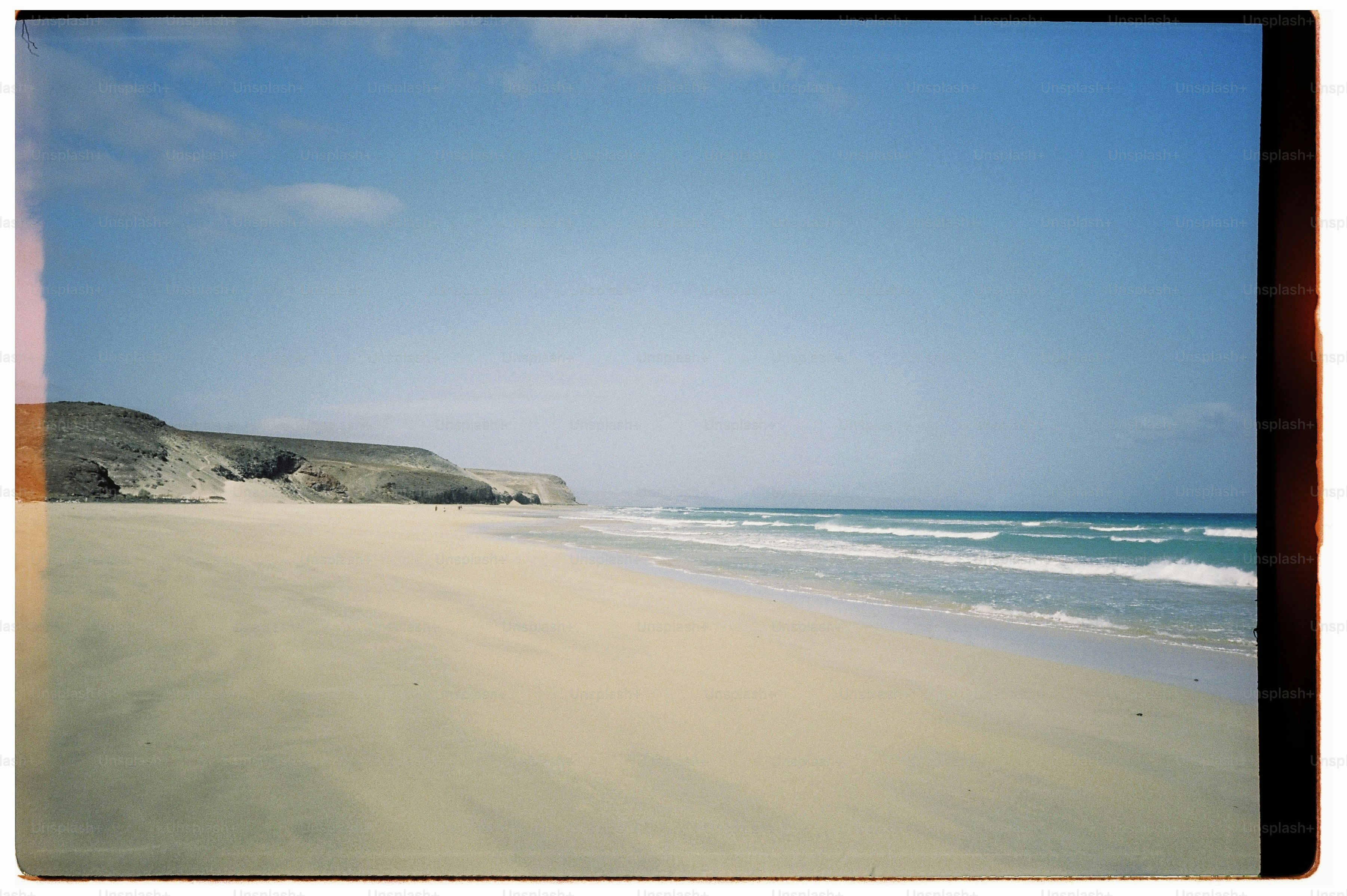 A wide sandy beach with turquoise waves meeting a rocky coastline under blue sky.