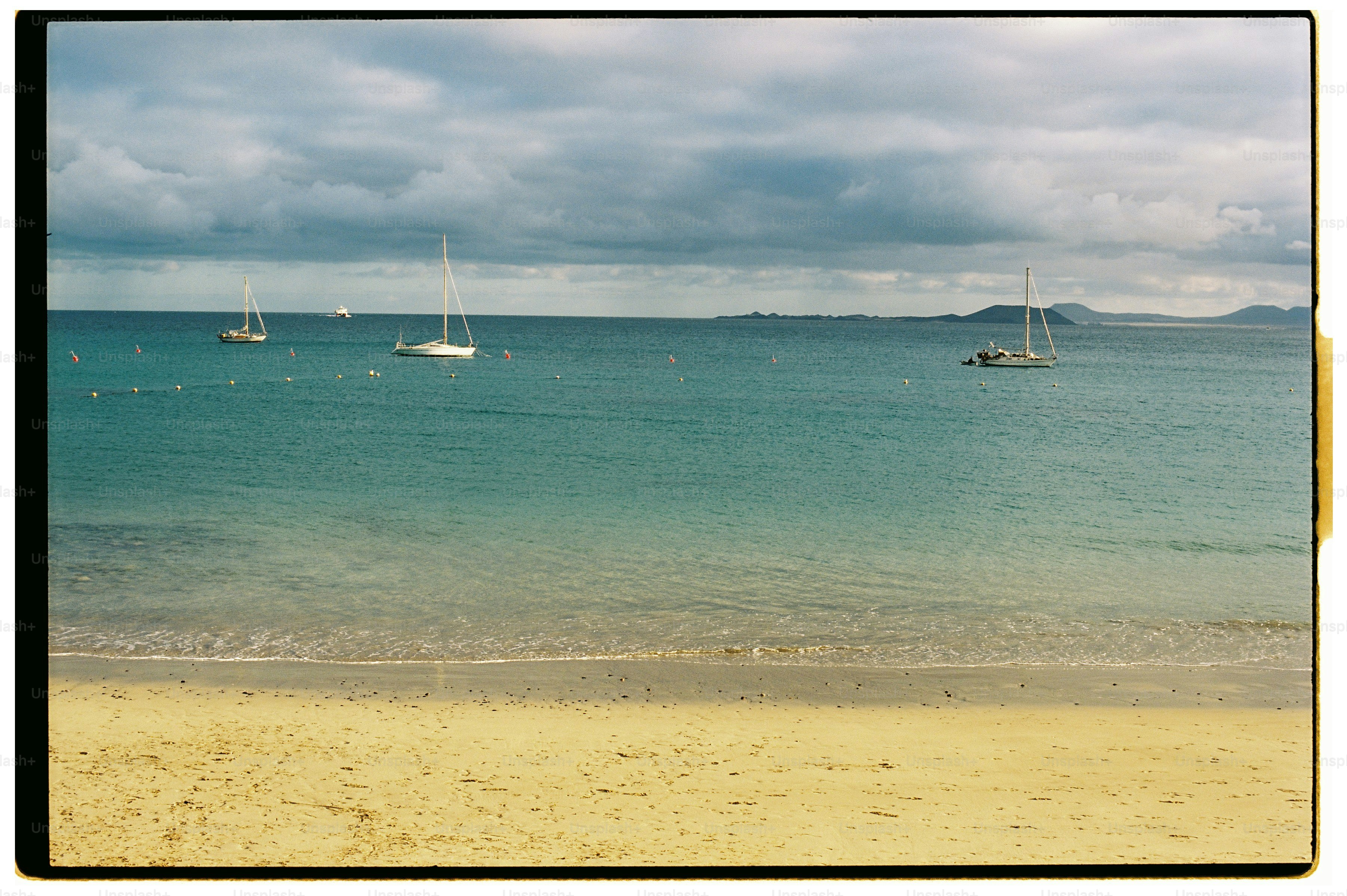 Three sailboats anchored in a calm turquoise bay under a cloudy sky.