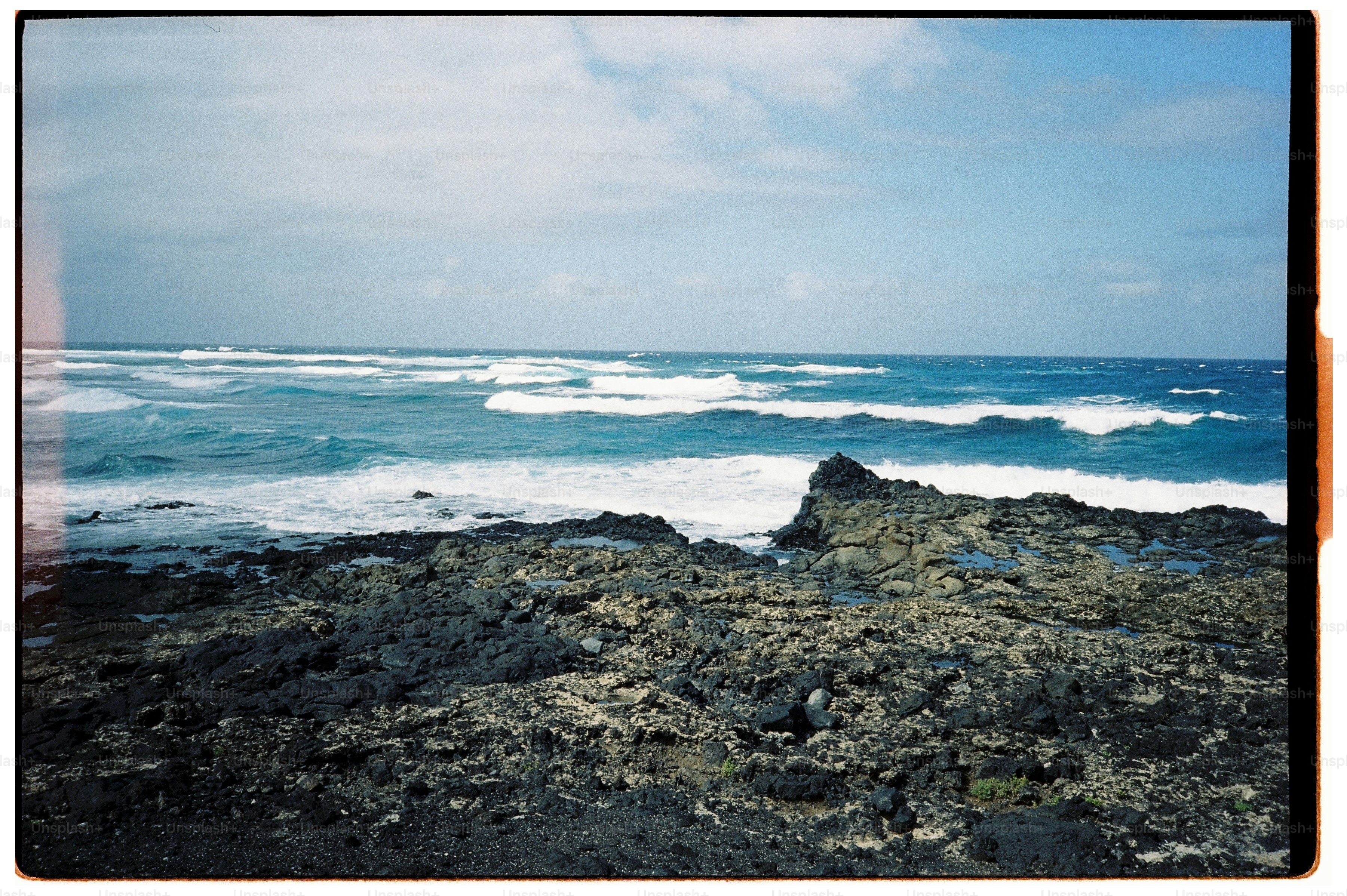 Waves crashing against a dark volcanic rock shoreline under a bright blue sky.