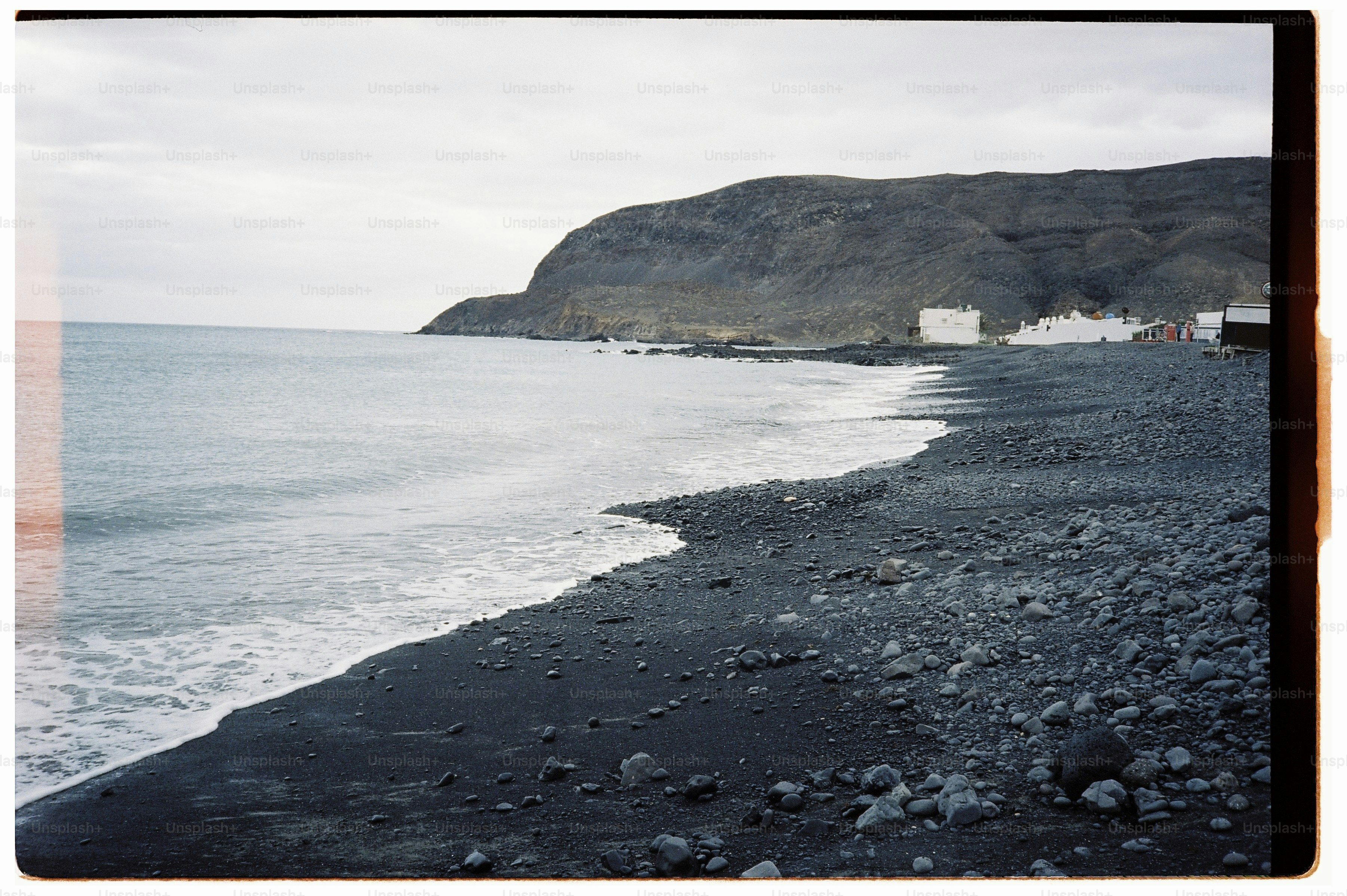 Waves washing onto a dark volcanic pebble beach beneath a large coastal cliff.