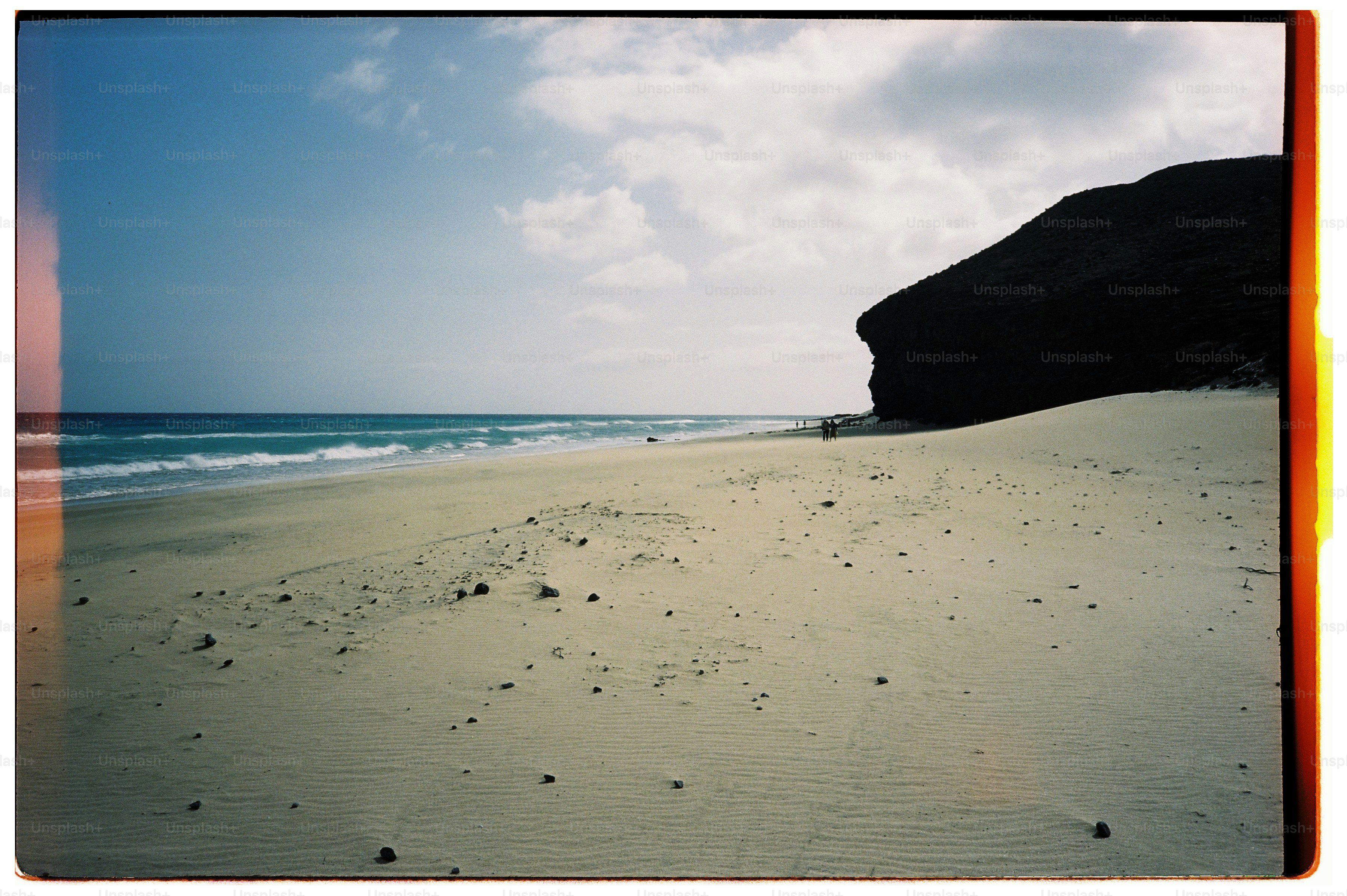 A sandy beach with a dark rocky cliff and distant people by the ocean.
