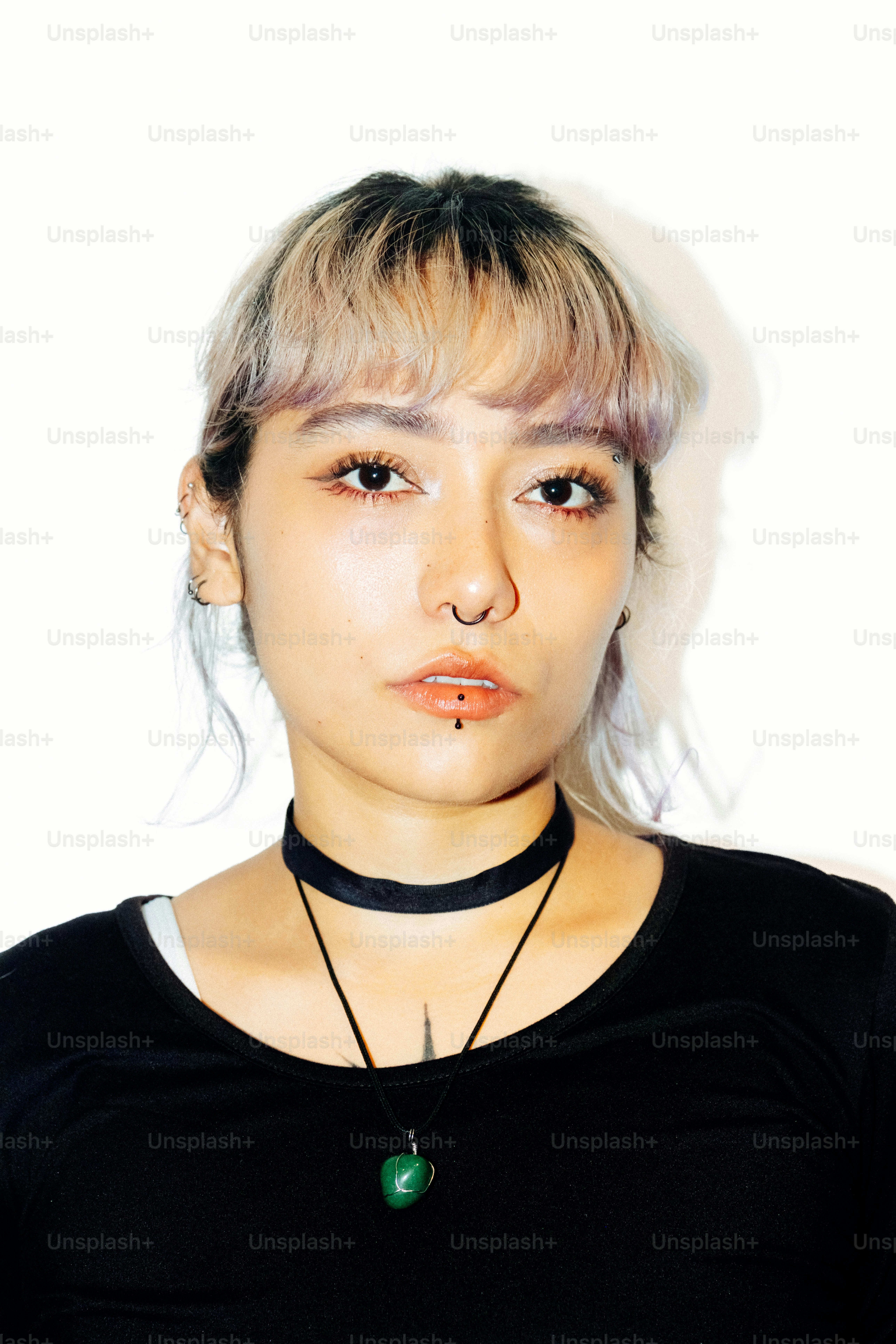 A person with blonde-streaked hair, facial piercings, and a green pendant necklace