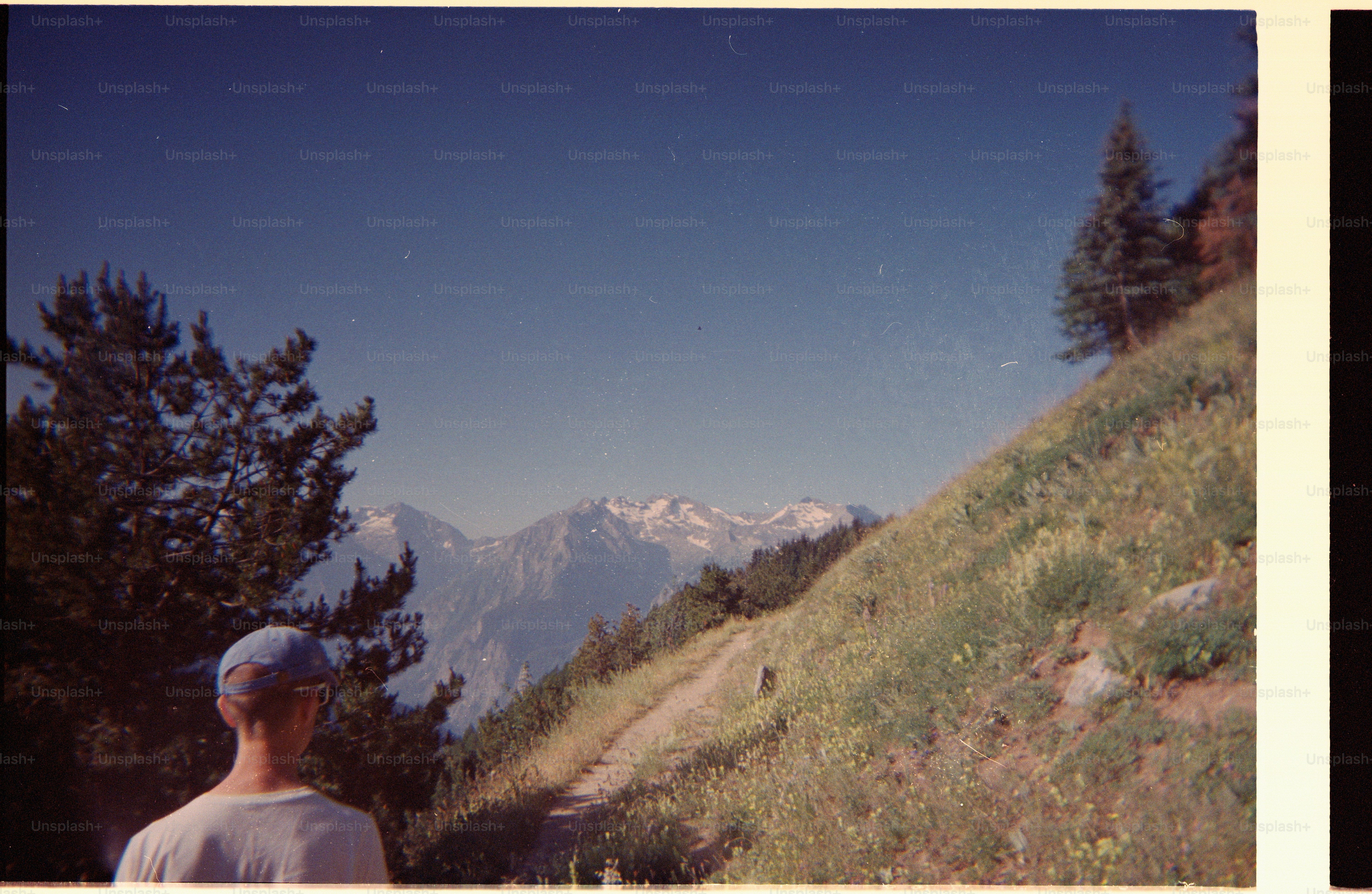 A person in a blue cap on a mountain trail overlooking distant snowy peaks.