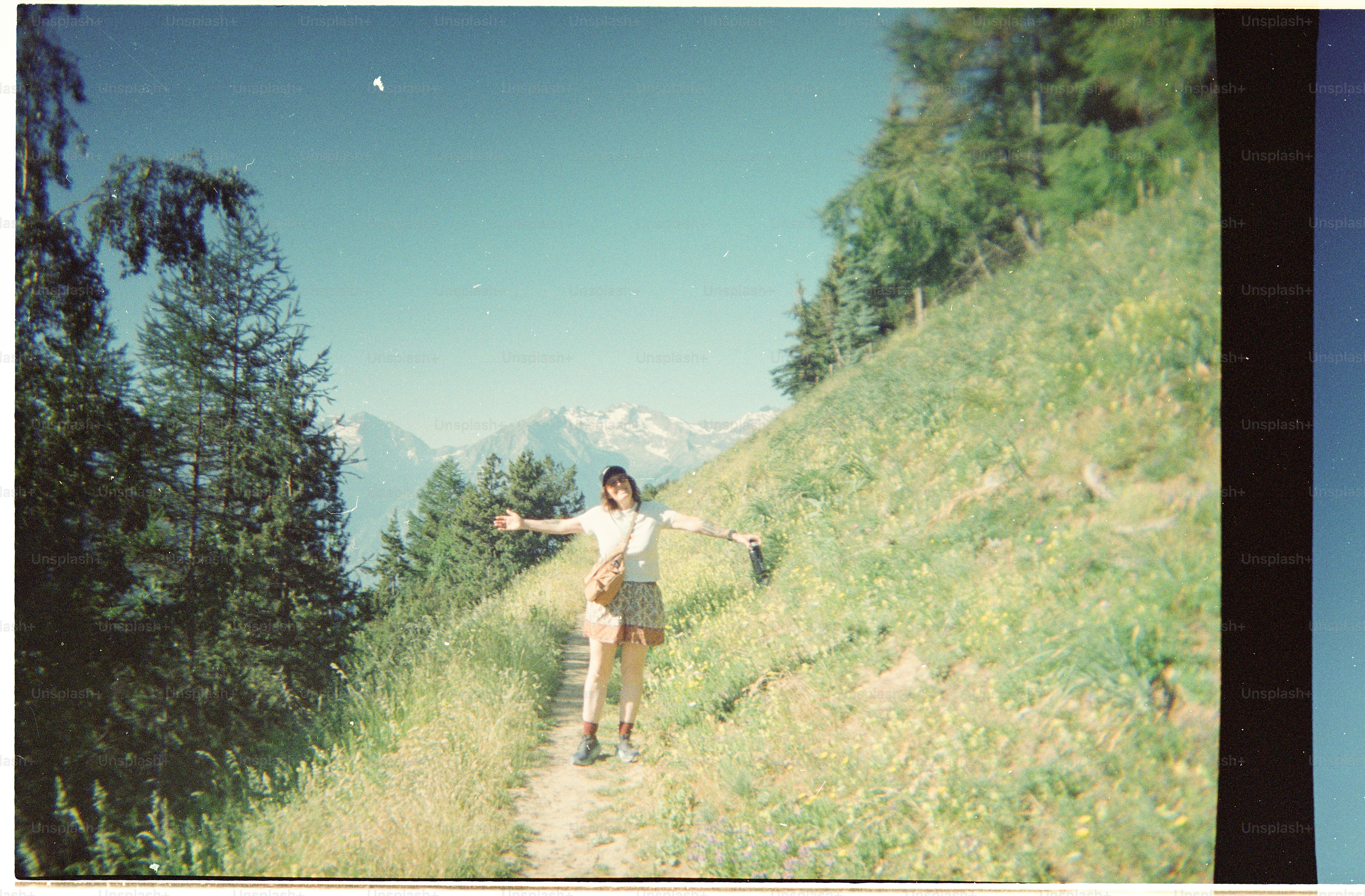 A person with arms outstretched on a mountain trail with pine trees and peaks.