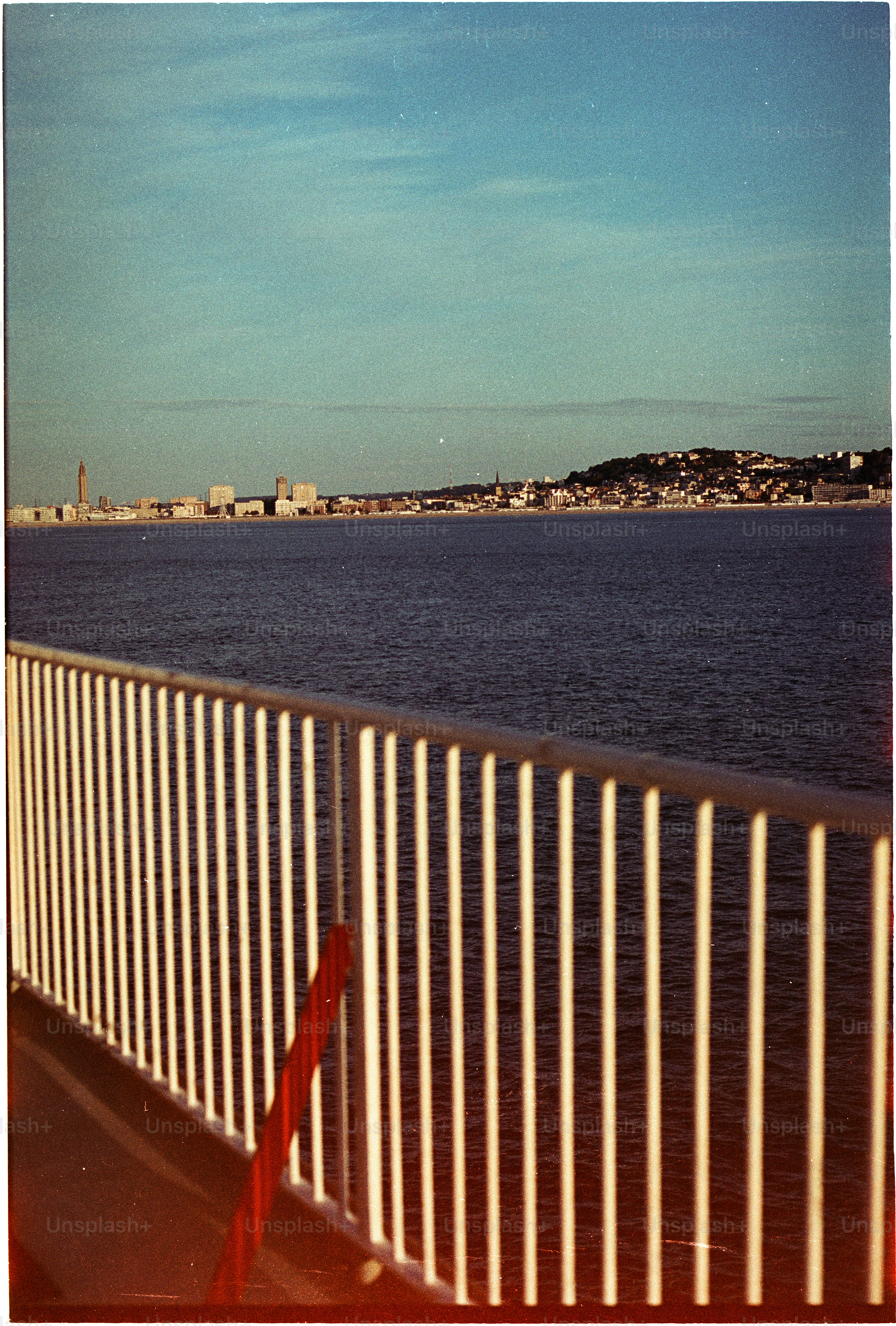 A white metal railing overlooking a calm sea with a distant coastal city skyline