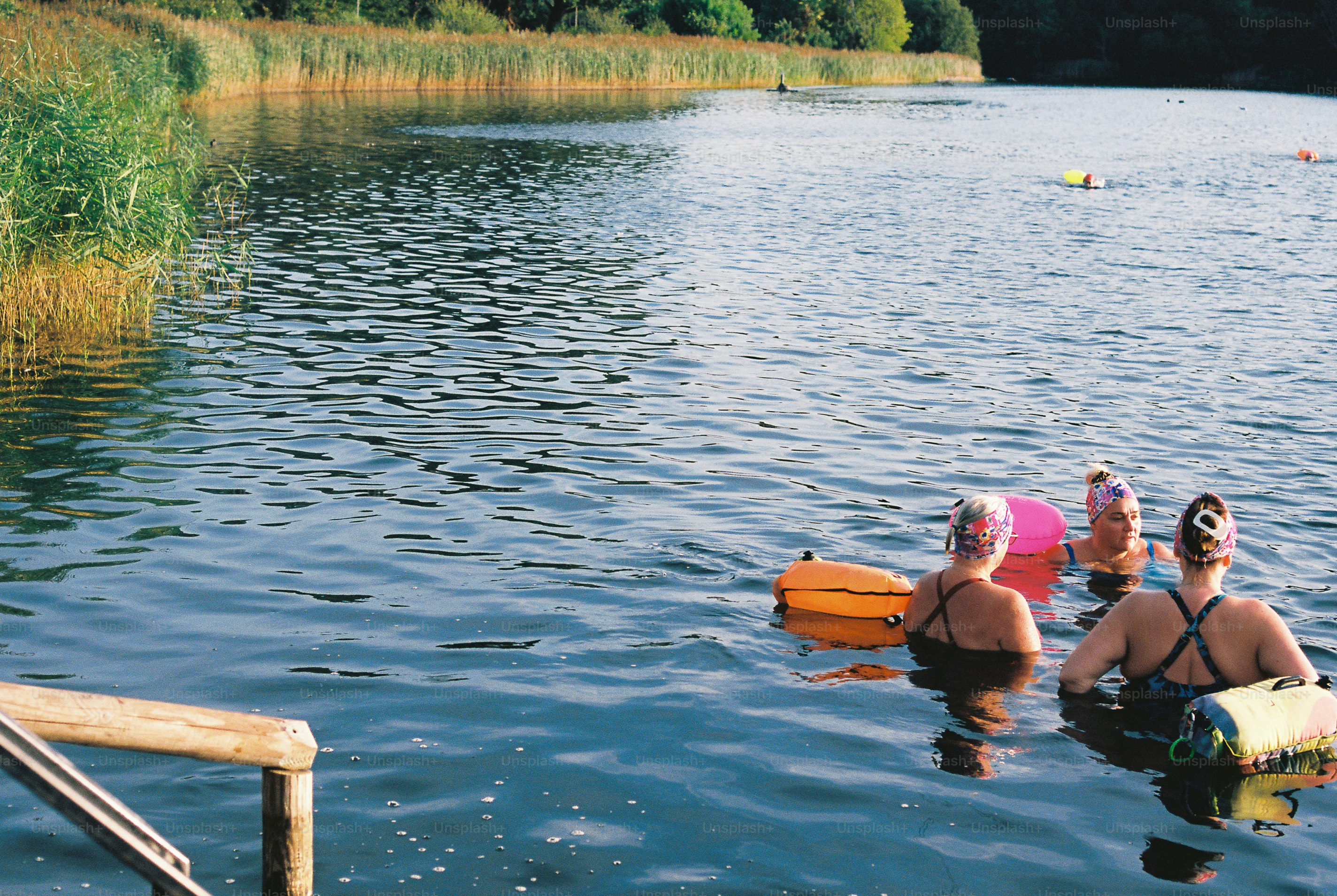 Three people swimming in a lake with colorful inflatable safety buoys.