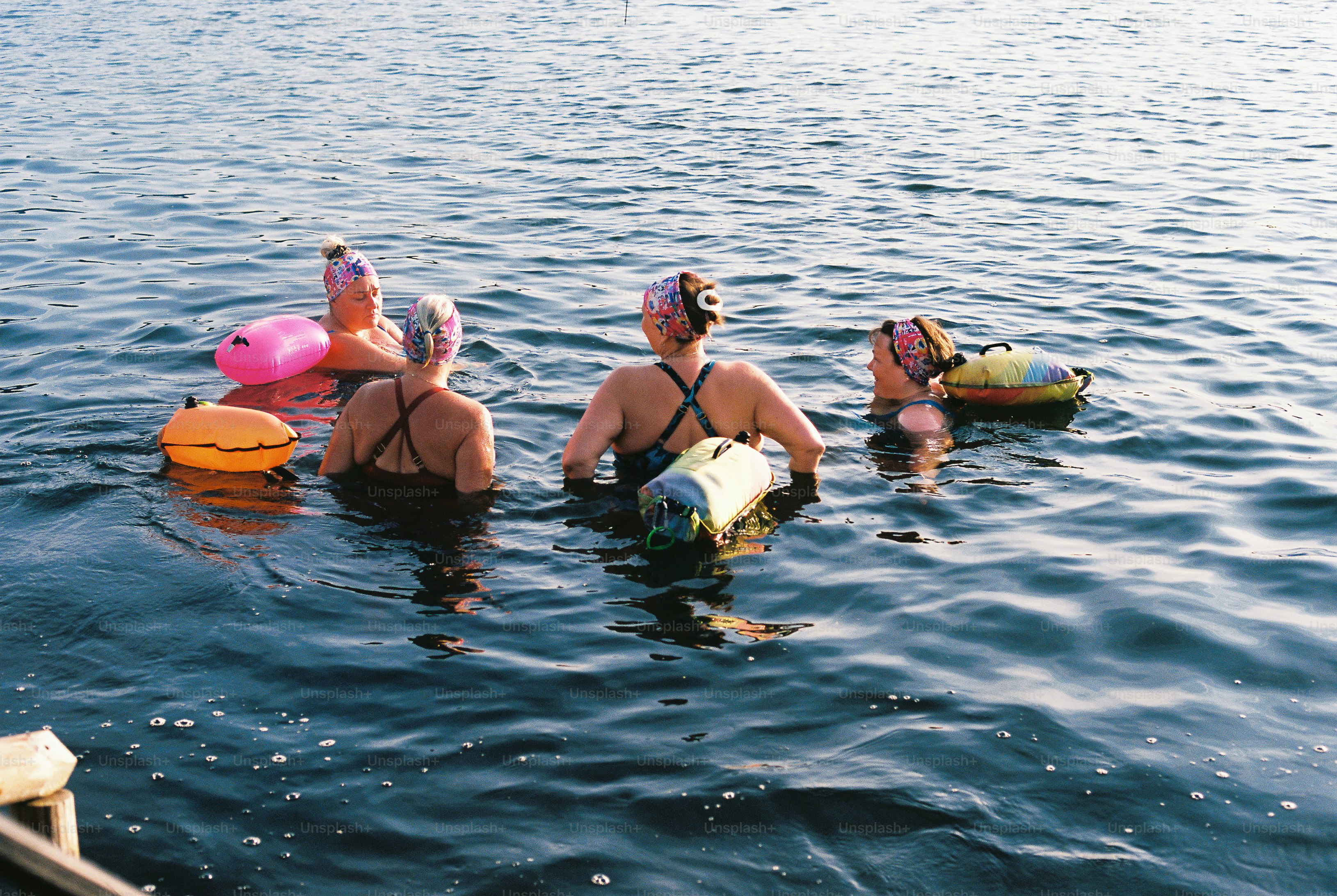 Four people in swim caps and swimwear floating in dark water with colorful dry bags.