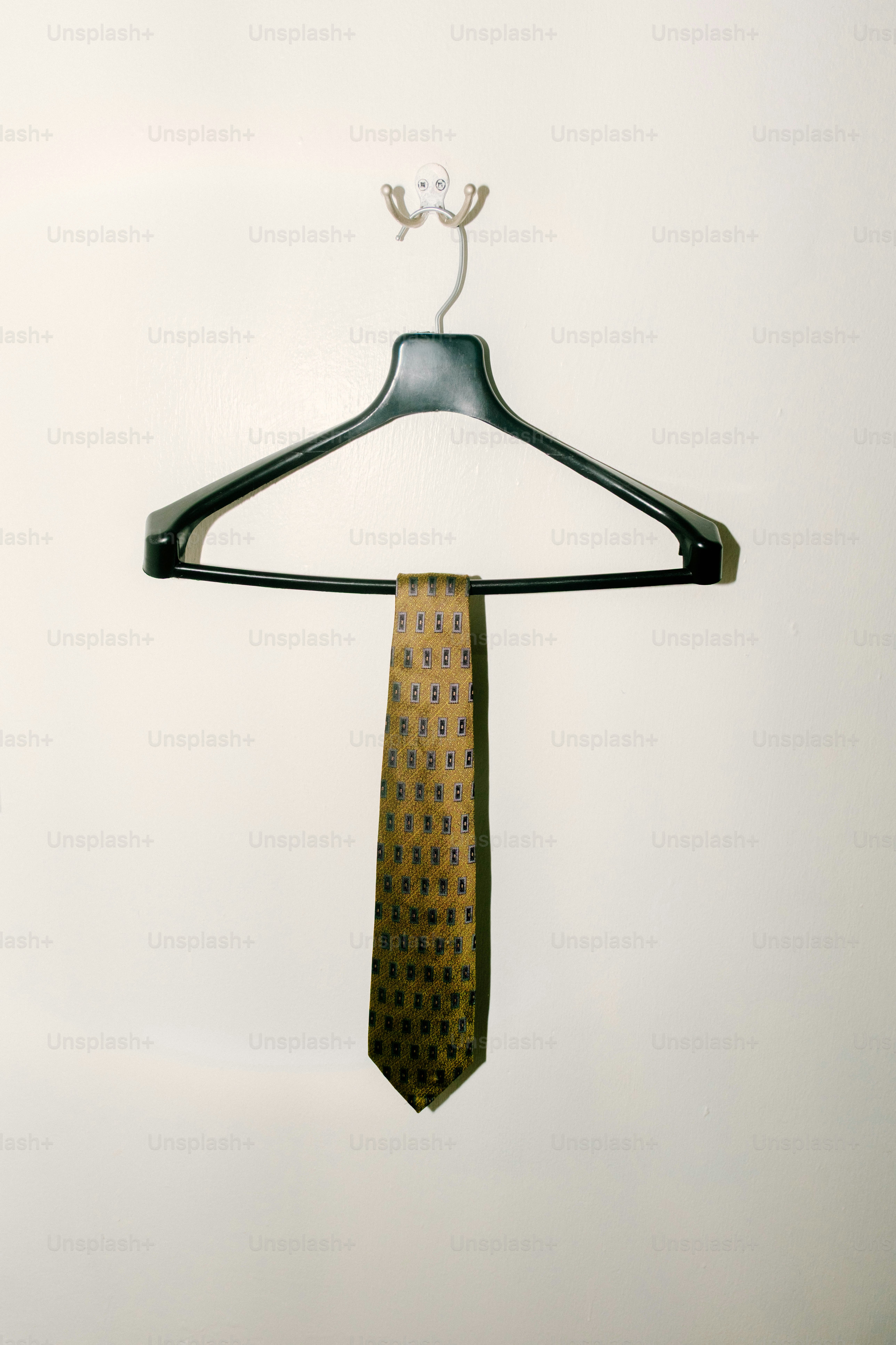 A patterned olive green necktie hanging from a black plastic hanger on a wall
