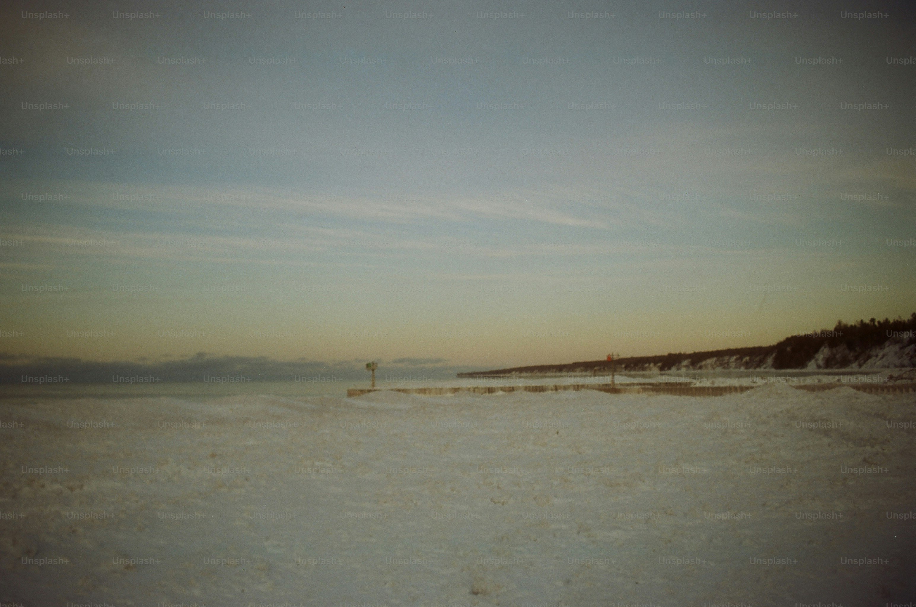 A snow and ice-covered lakeshore stretches beneath a pastel dusk sky, with a frozen pier and wooded bluff visible in the distance. The scene evokes quiet winter solitude.