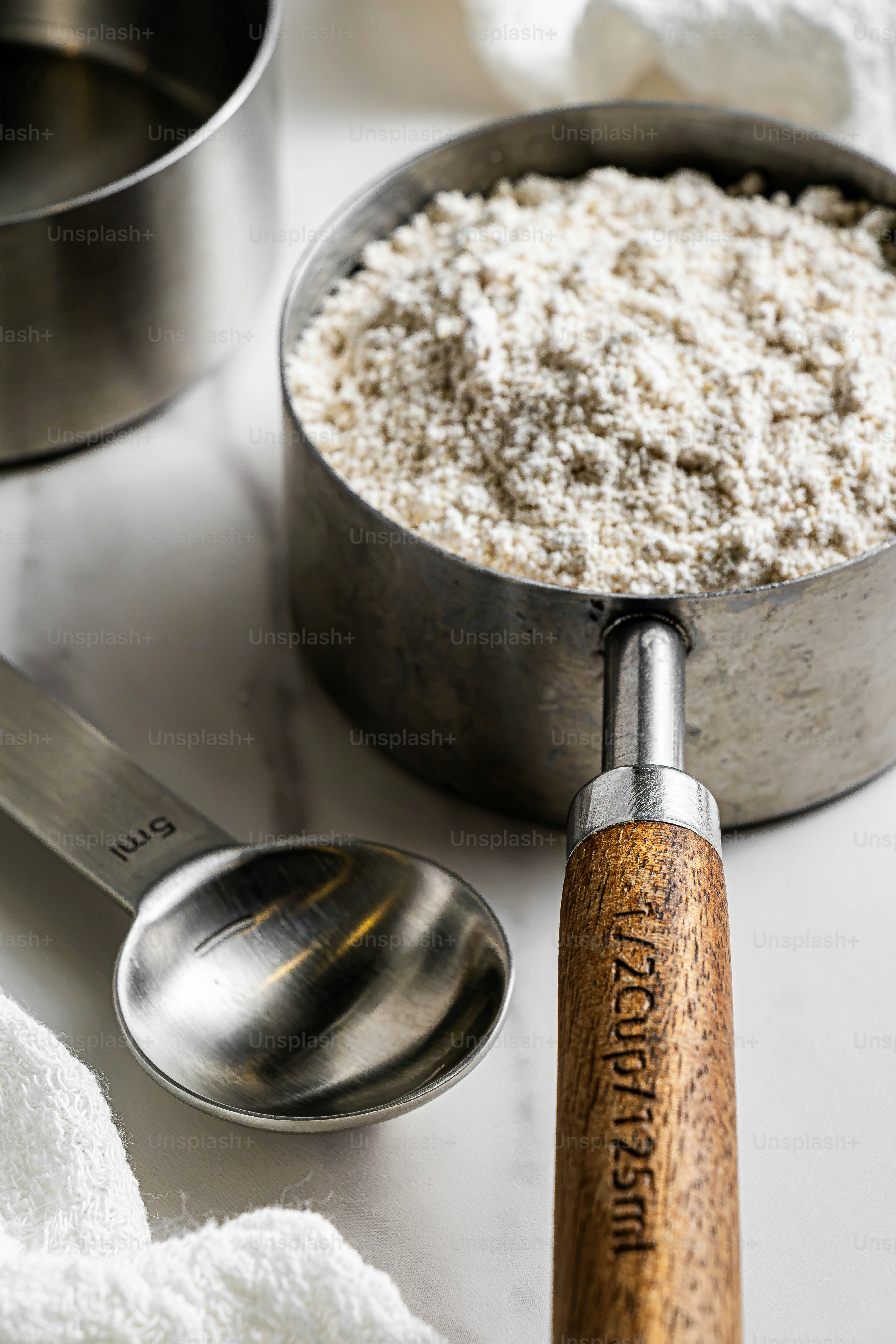 A metal measuring cup filled with flour beside a small metal spoon