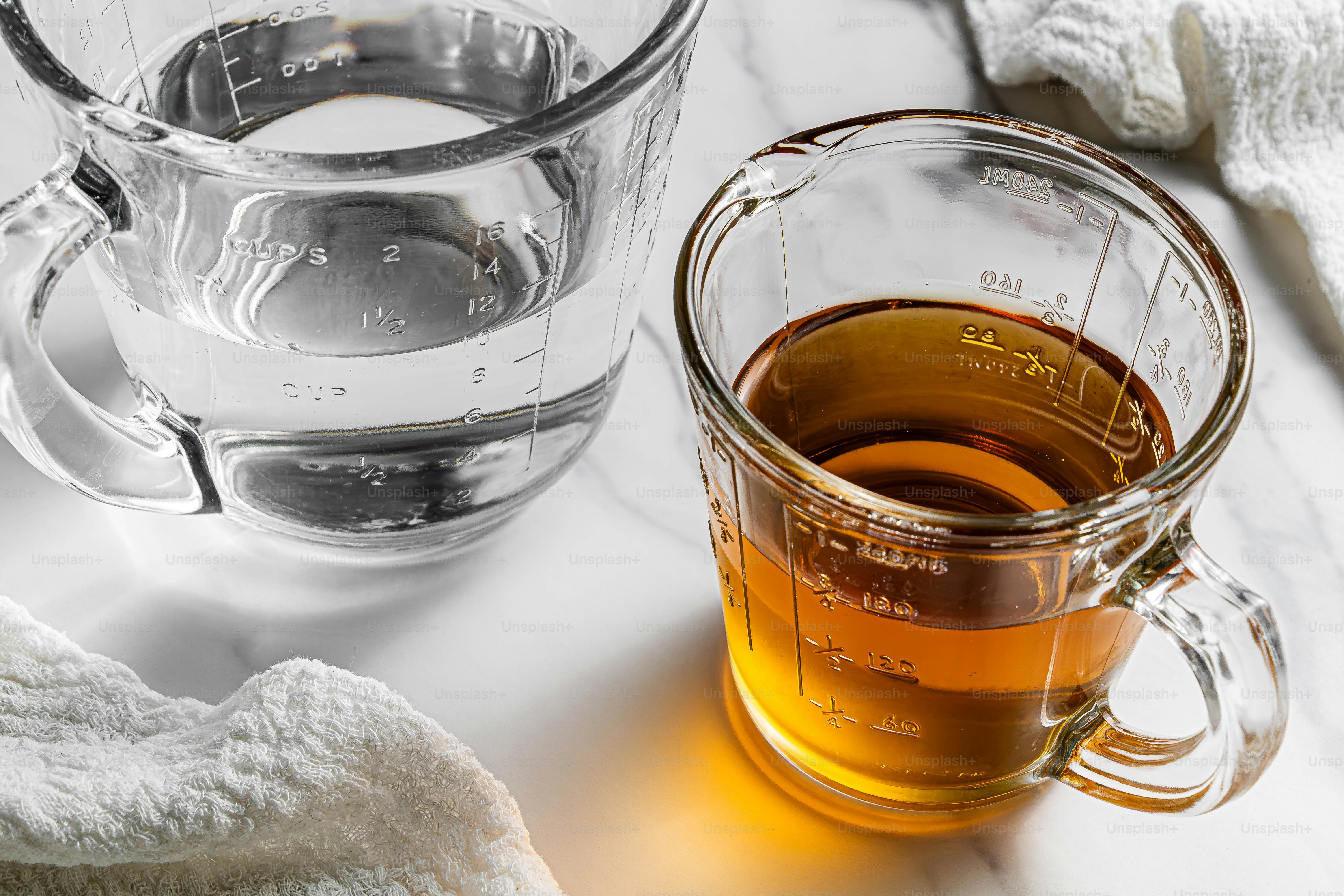 Two glass measuring cups containing clear water and amber liquid on a marble surface