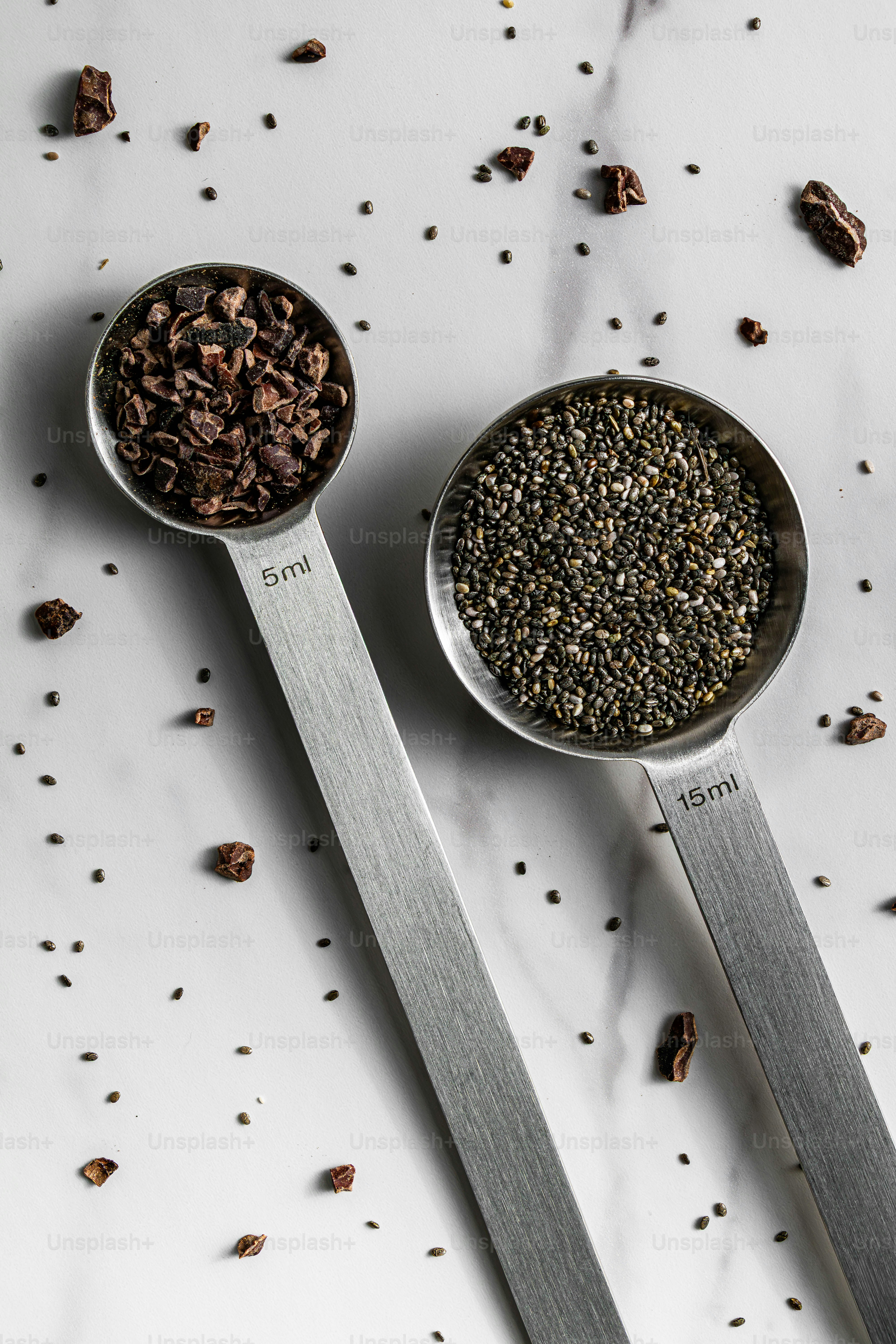 Cacao nibs and chia seeds in two metal measuring spoons on a white surface