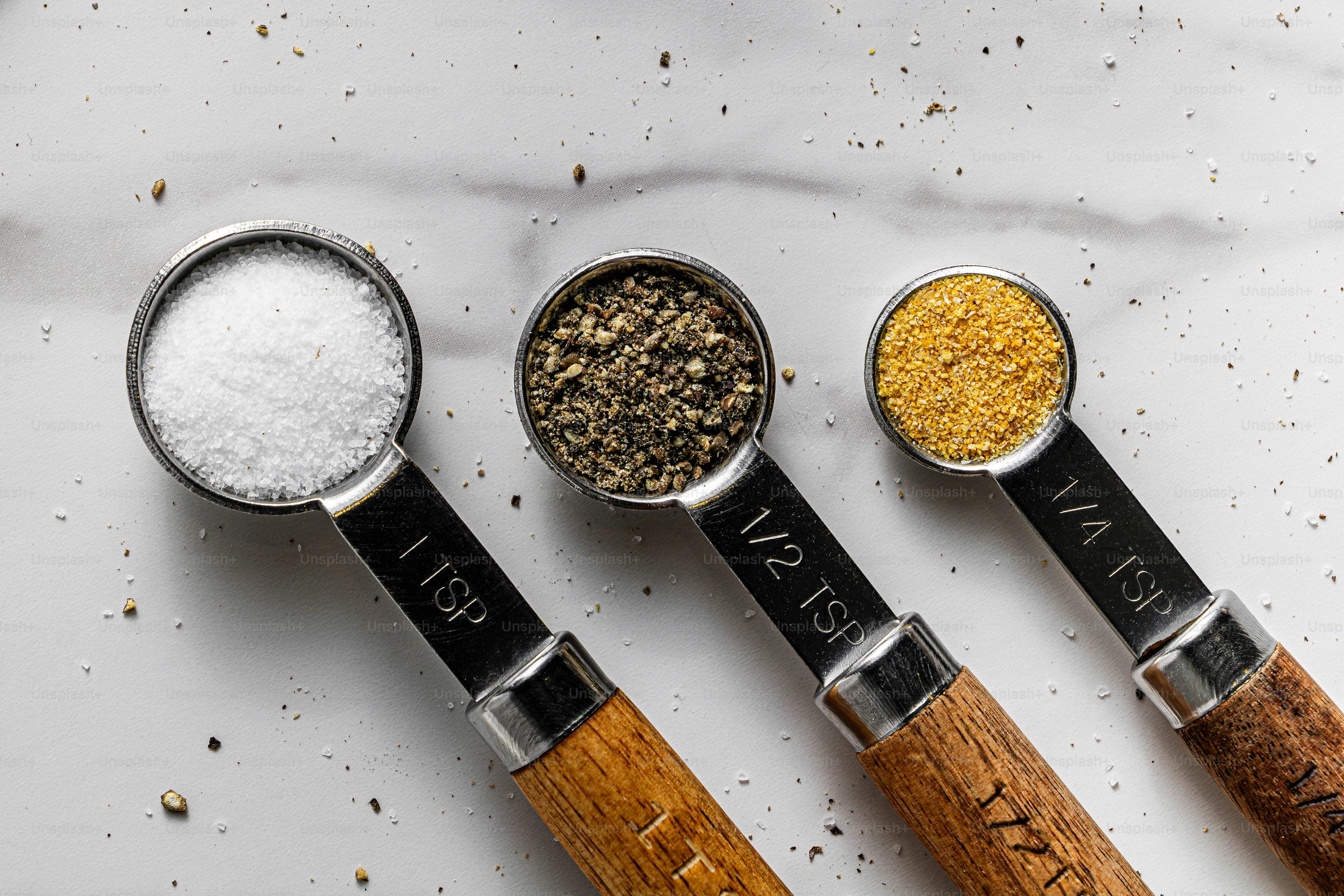 Three measuring spoons filled with salt, black pepper, and yellow spice on marble